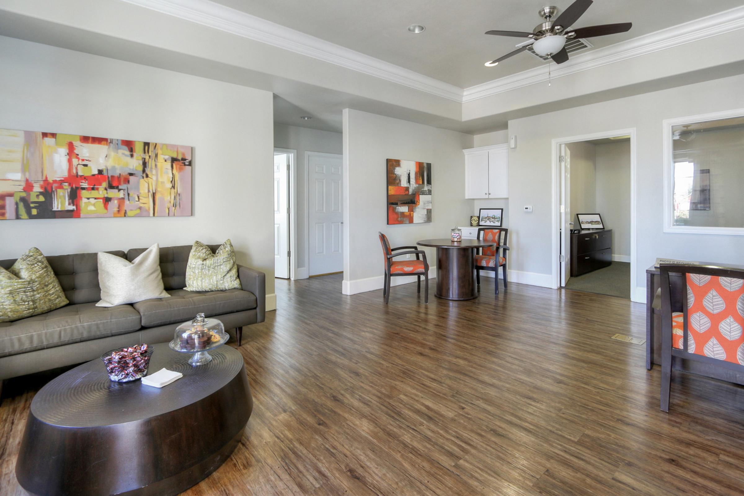A modern living area featuring a comfortable gray sofa with decorative pillows, a round wooden coffee table, and vibrant artwork on the walls. A dining area with four chairs is visible, and there's a light-colored door leading to another room. The floor has a warm wooden finish, creating a cozy atmosphere.
