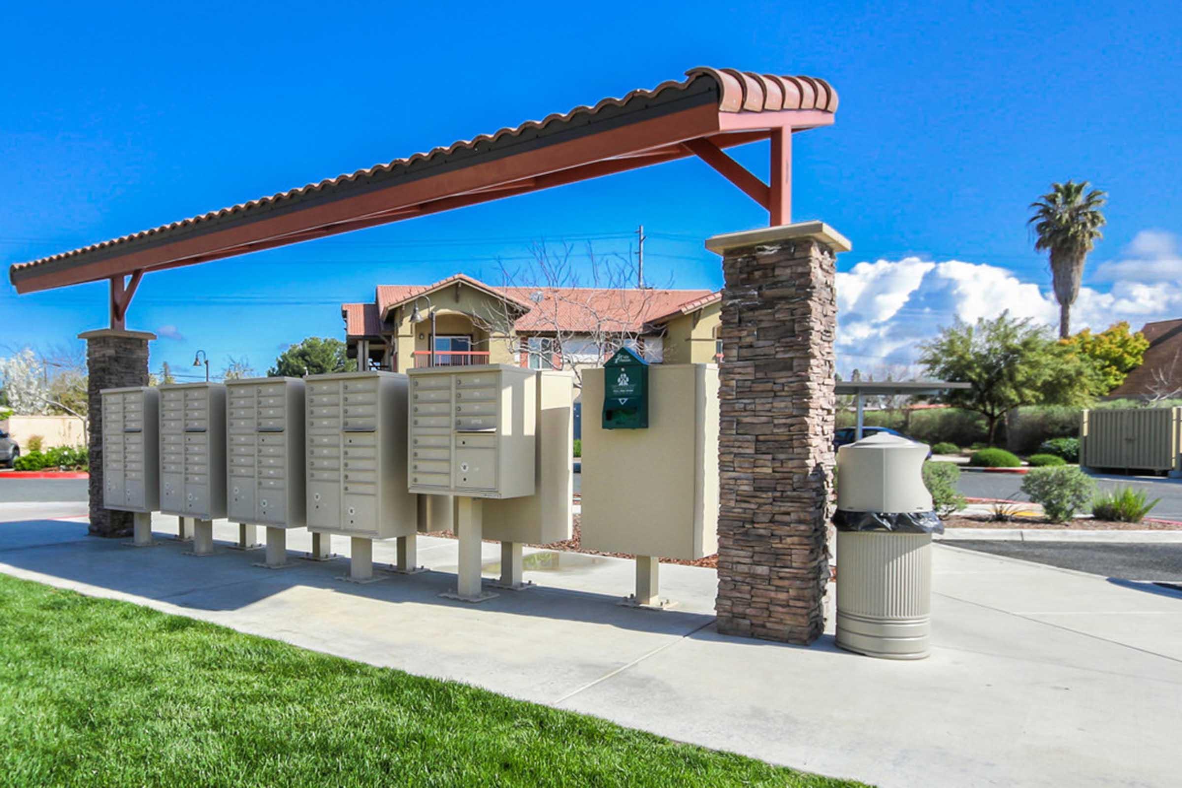 Multiple mailboxes installed in a row under a pergola, with a waste bin nearby, set against a bright blue sky and a green lawn. In the background, there are residential buildings and palm trees, indicating a well-maintained community area.