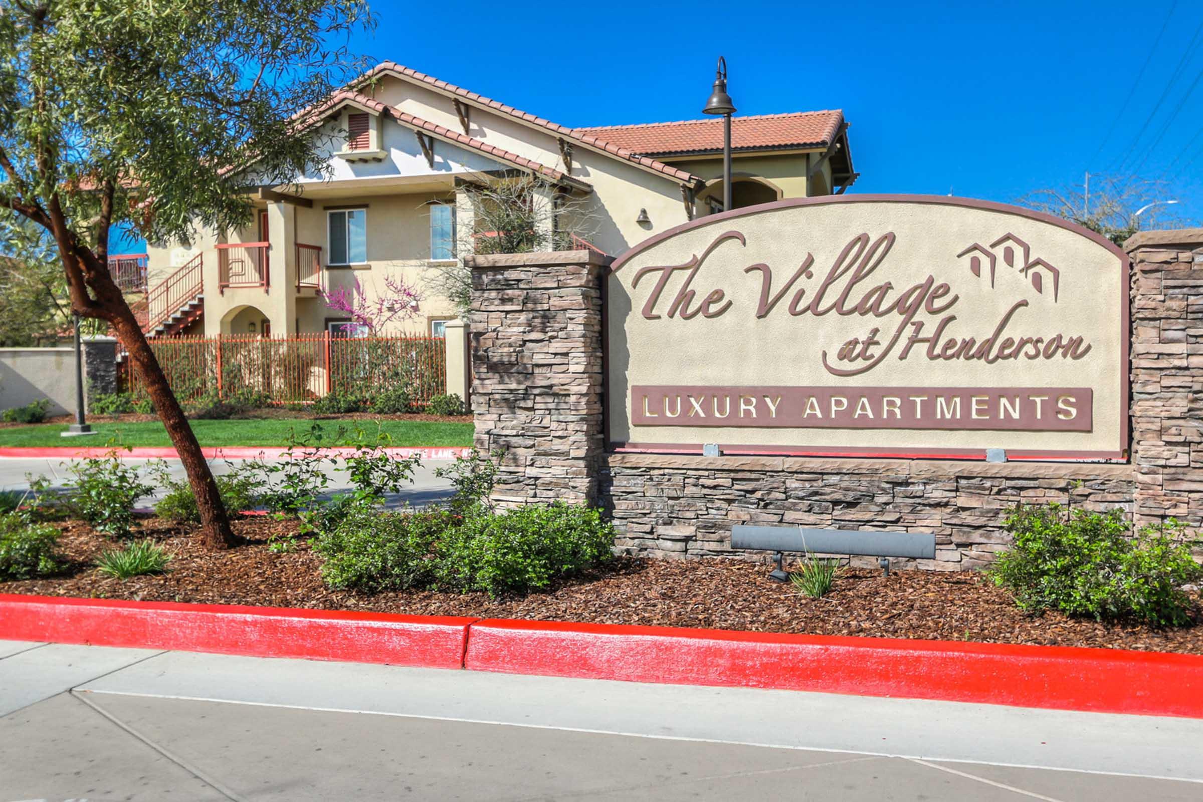 Sign for "The Village at Henderson Luxury Apartments" with a stone base, surrounded by landscaped greenery and trees. In the background, a multi-story apartment building with a tiled roof and clear blue sky. The pathway is paved, leading towards the entrance of the complex.