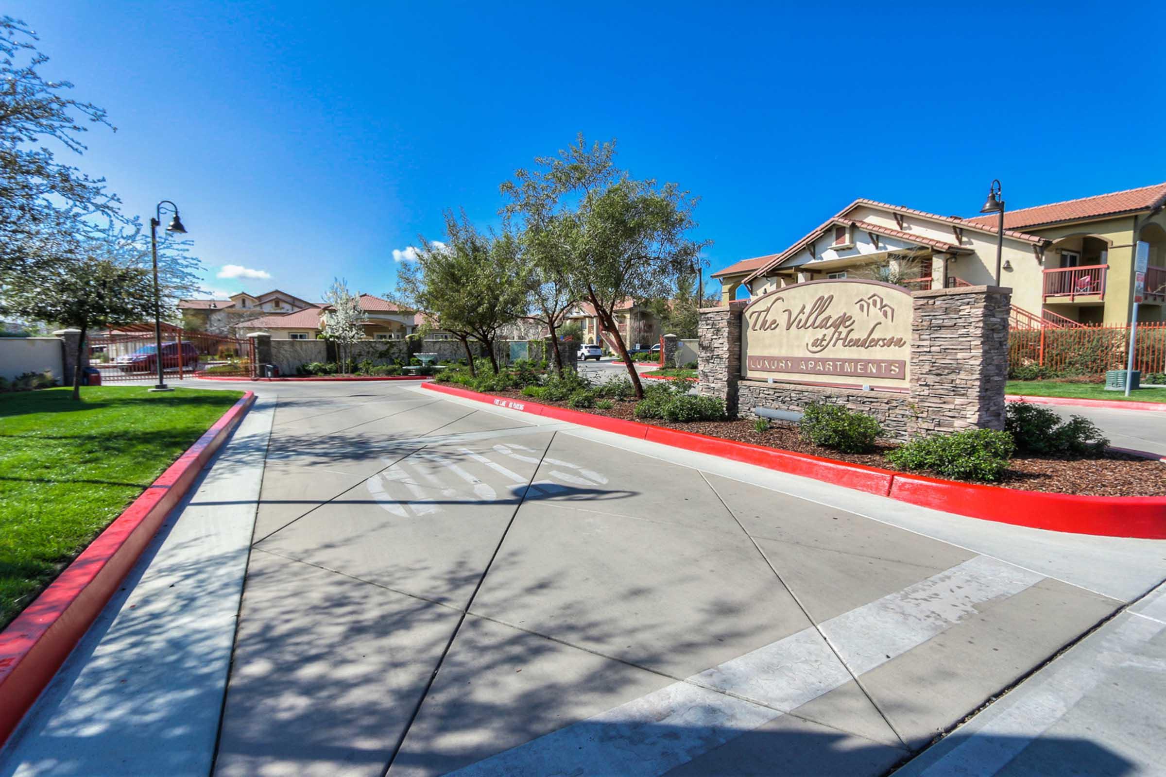 Sign for "The Valley at Hudson" apartment complex, with a landscaped pathway and trees on either side. The entrance features a modern design and clear blue sky in the background, indicating a sunny day.
