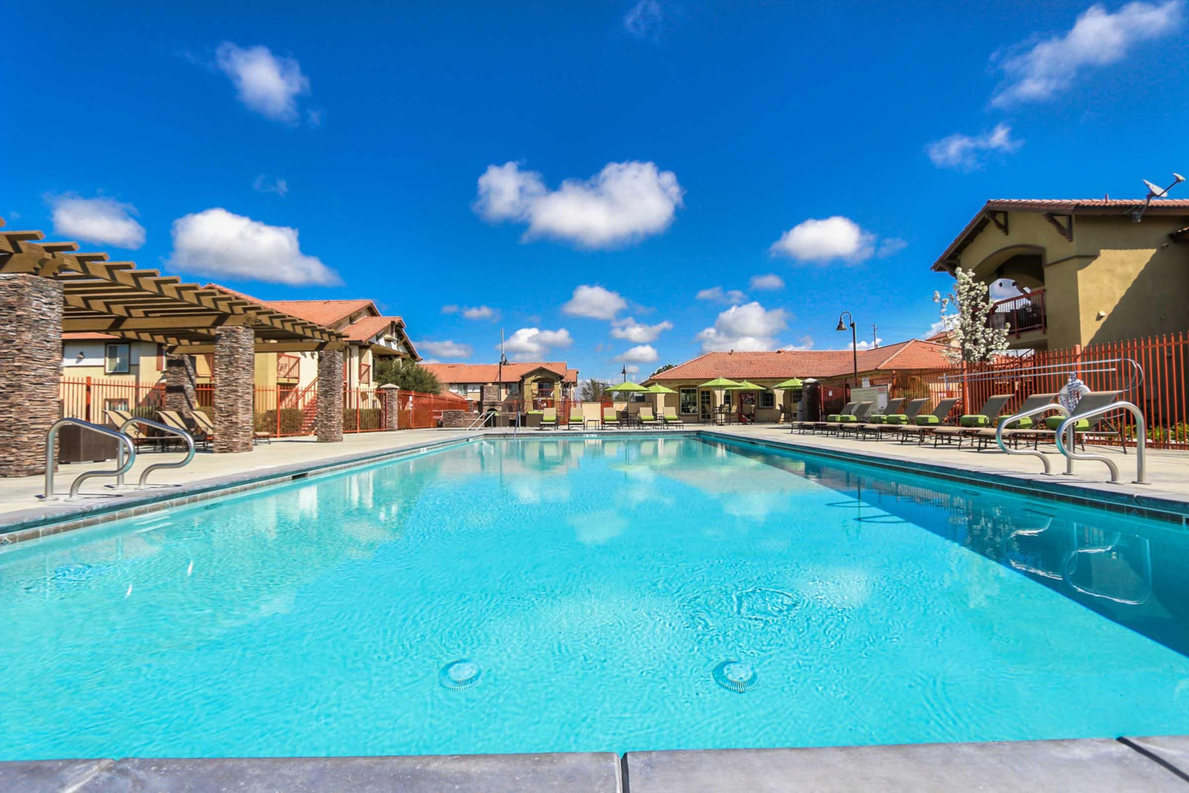 A clear swimming pool surrounded by lounge chairs and shaded areas, with a backdrop of residential buildings under a bright blue sky with fluffy white clouds. The scene conveys a resort-like atmosphere, perfect for relaxation and leisure.
