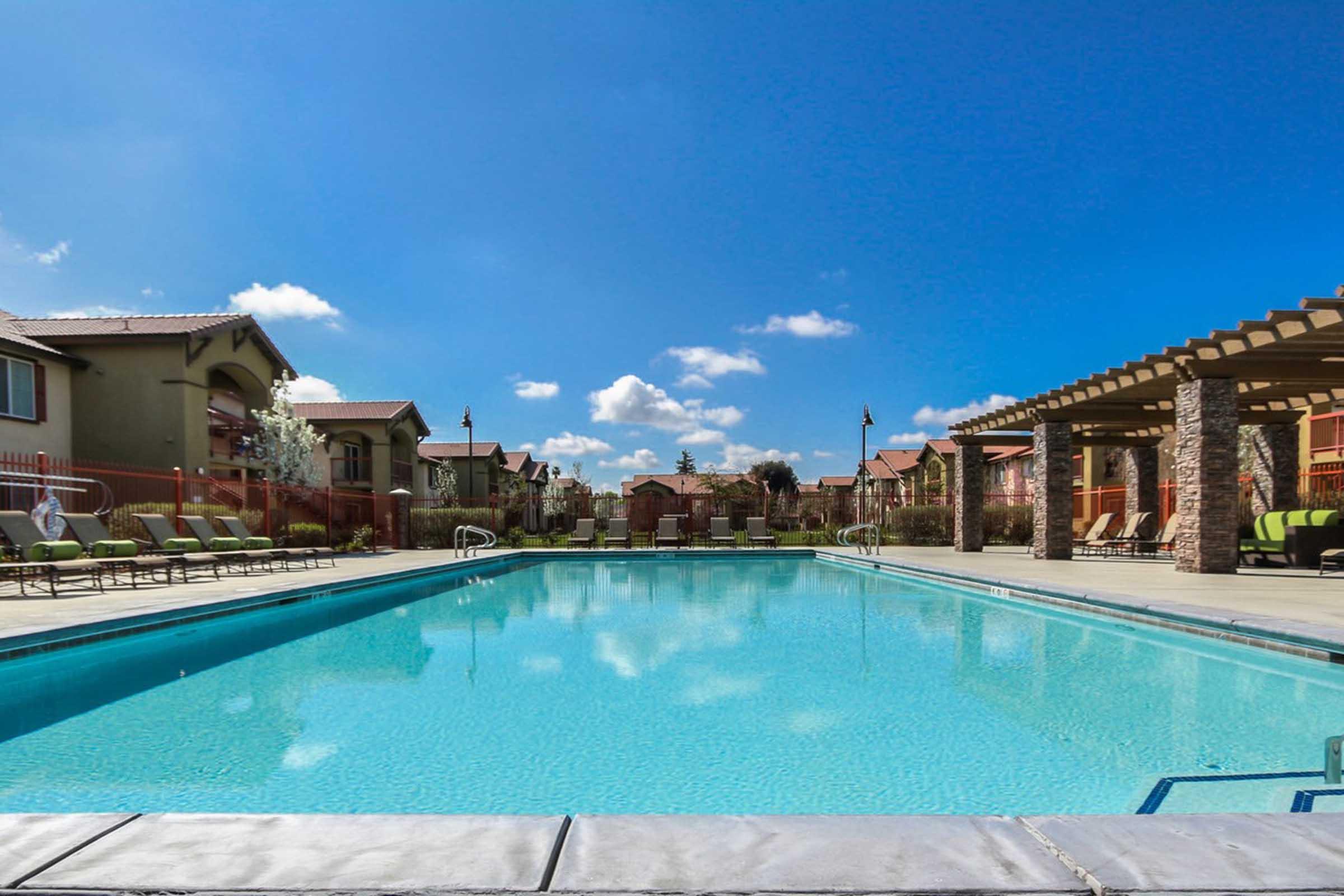 A clear swimming pool surrounded by lounge chairs, with residential buildings and greenery in the background. The sky is blue with a few clouds, creating a serene and inviting atmosphere.