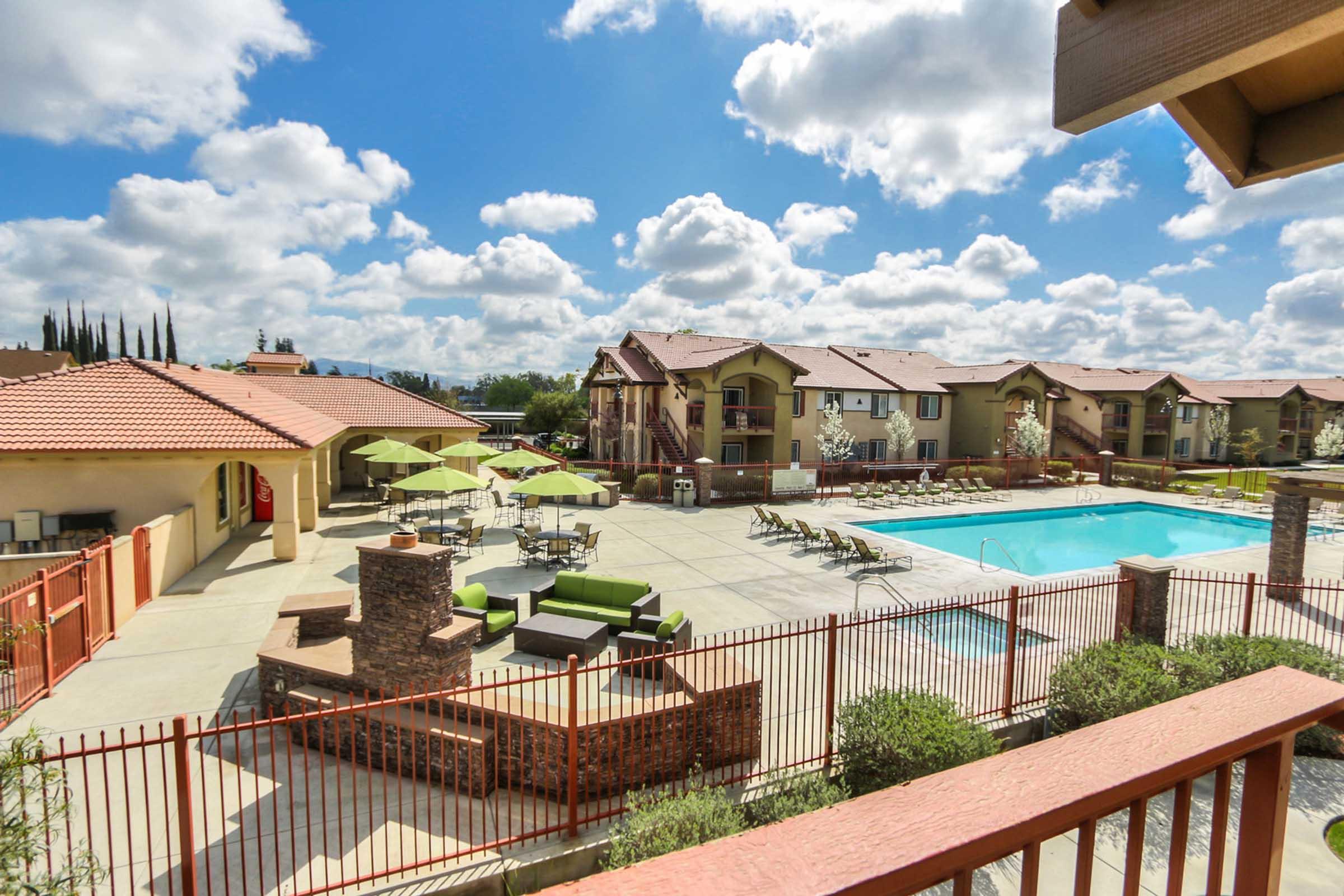 A view of a sunny apartment complex featuring a large swimming pool surrounded by lounge chairs and green seating areas. Umbrellas provide shade for outdoor tables. In the background, there are several two-story buildings and a clear blue sky with fluffy clouds.