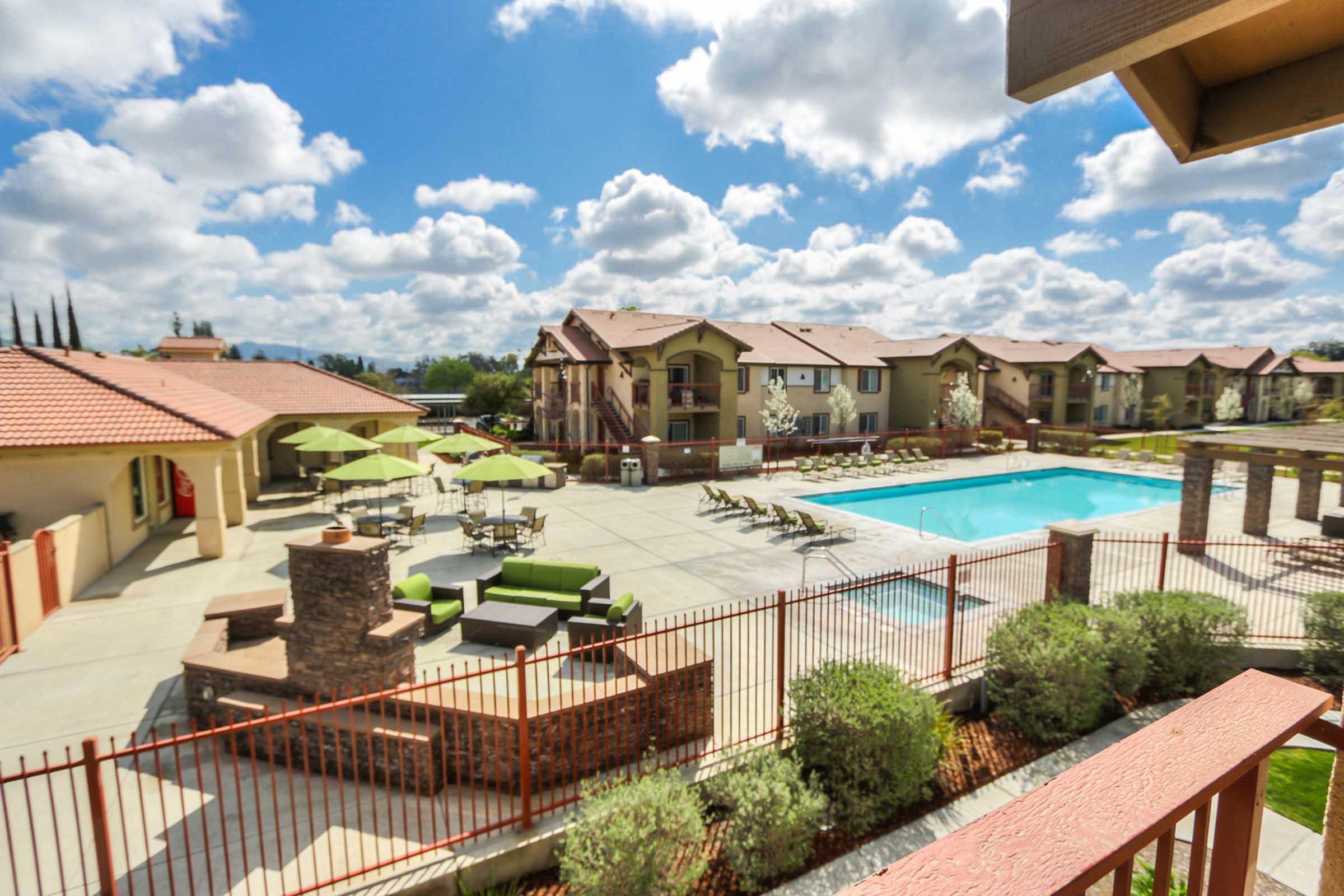 A spacious outdoor area featuring a swimming pool surrounded by lounge chairs, green umbrellas, and modern seating. Nearby, there are residential buildings with terracotta roofs and well-maintained landscaping. The sky is partly cloudy, adding to the inviting atmosphere.