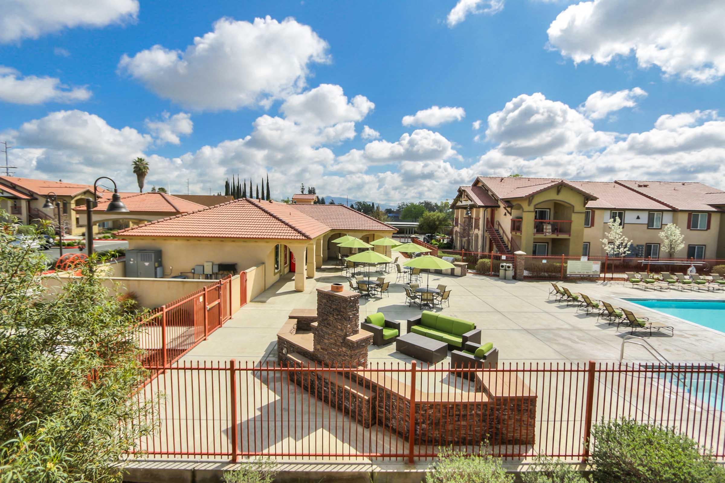 A sunny outdoor area featuring a swimming pool surrounded by lounge chairs. There are several green umbrellas and patio furniture, including a seating area with a stone fireplace. In the background, there are residential buildings and a clear blue sky with fluffy clouds.