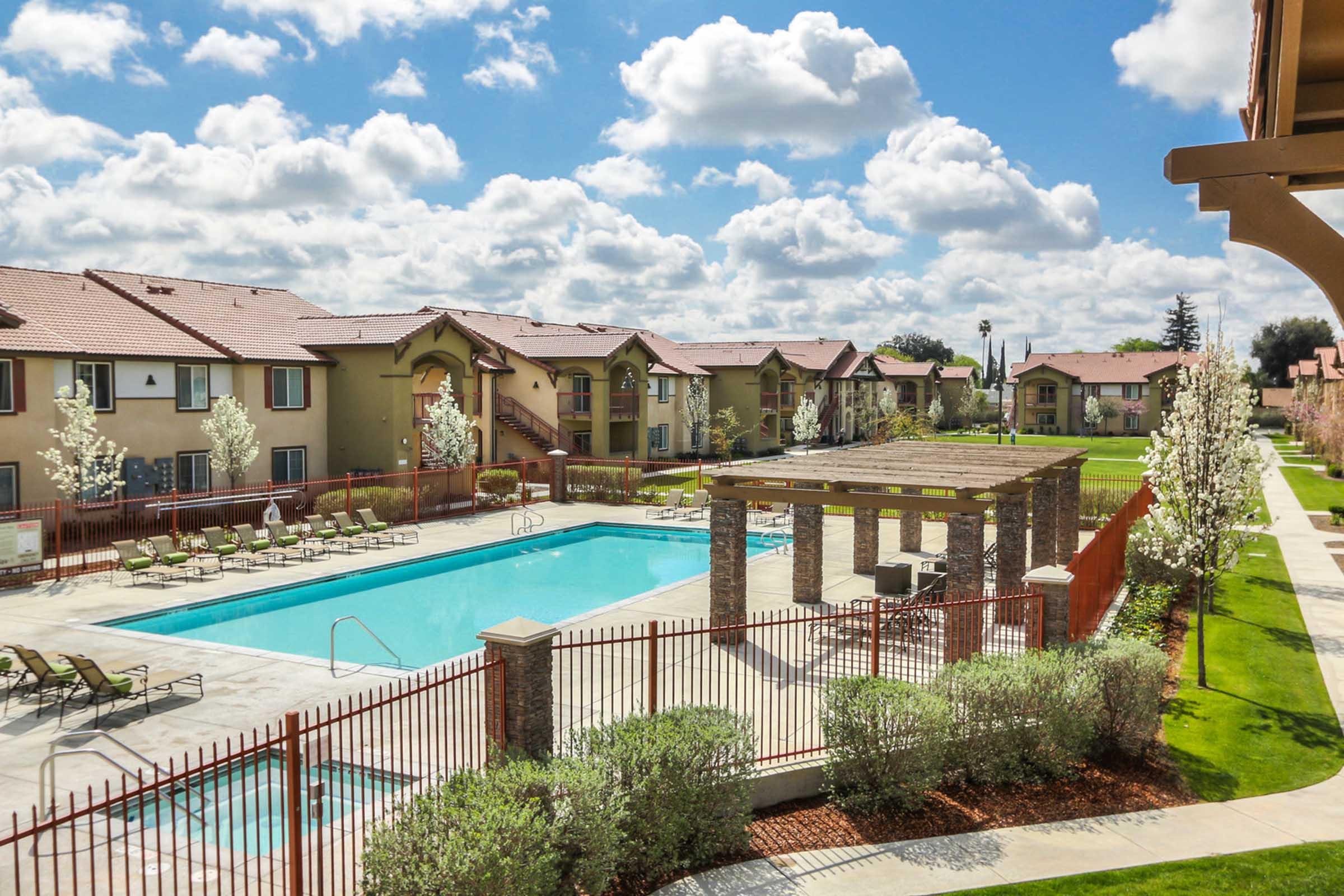 A bright and inviting view of a residential complex featuring a swimming pool surrounded by lounge chairs. The area is landscaped with trees and shrubs, and there are several two-story apartment buildings in the background under a partly cloudy sky.