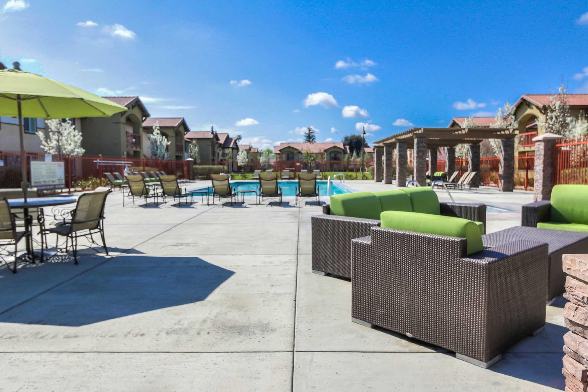 A sunny outdoor pool area featuring a clear blue sky, comfortable green and brown lounge seating, dining tables, and a well-maintained pool. Surrounding the pool are homes and landscaped areas with trees and shrubs, creating a relaxing atmosphere.