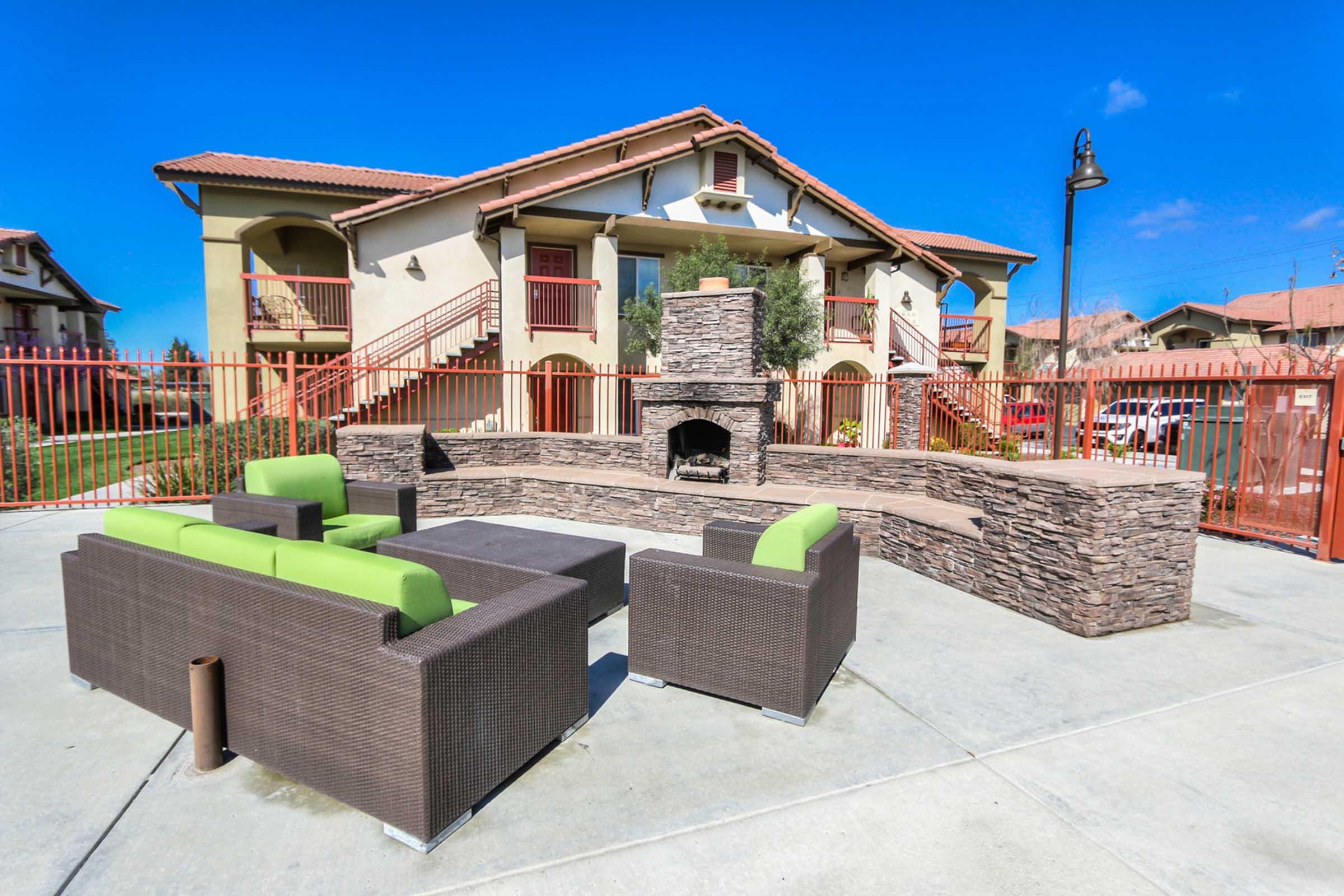 A cozy outdoor seating area featuring modern green and dark brown furniture around a stone fireplace, set against a backdrop of a two-story apartment building with red balconies and a clear blue sky. The space is enclosed by a red fence, creating a private and inviting atmosphere.