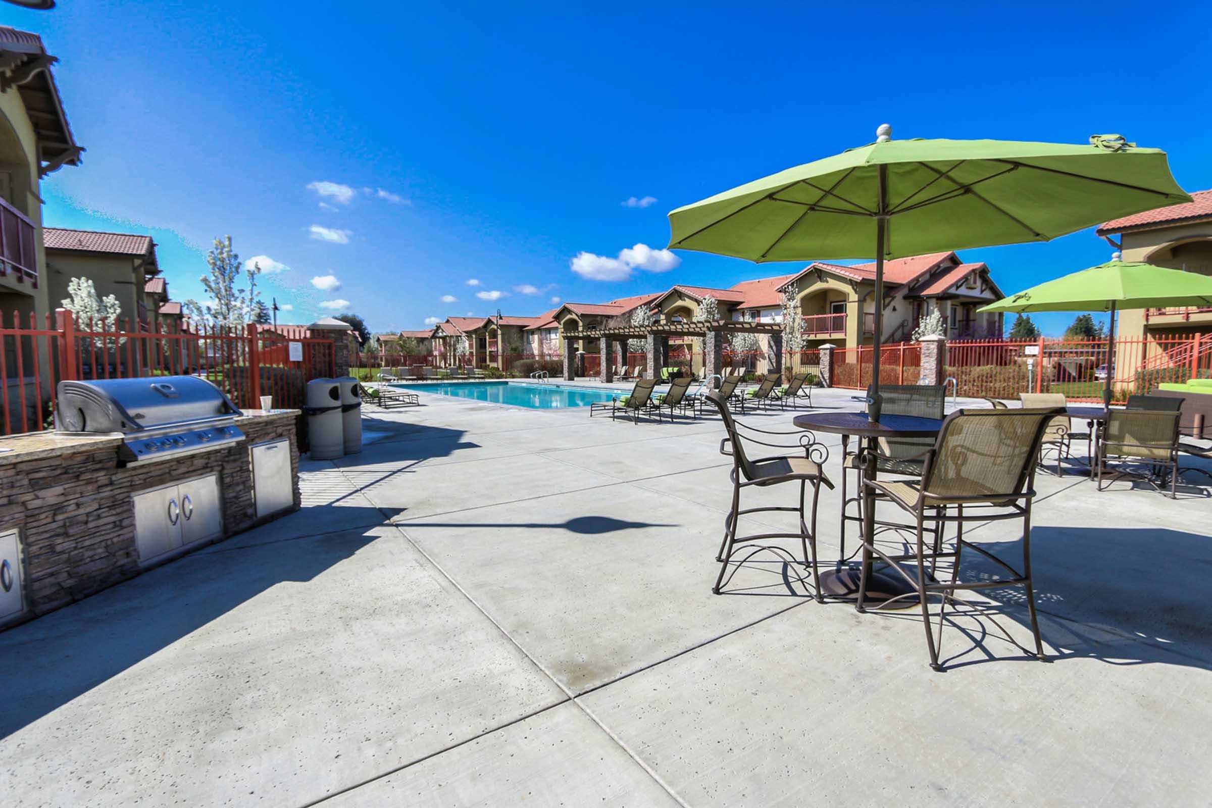A sunny pool area featuring lounge chairs and green umbrellas, surrounded by residential buildings. There's a barbecue grill nearby, and a clear blue sky adds to the inviting atmosphere of the outdoor space.