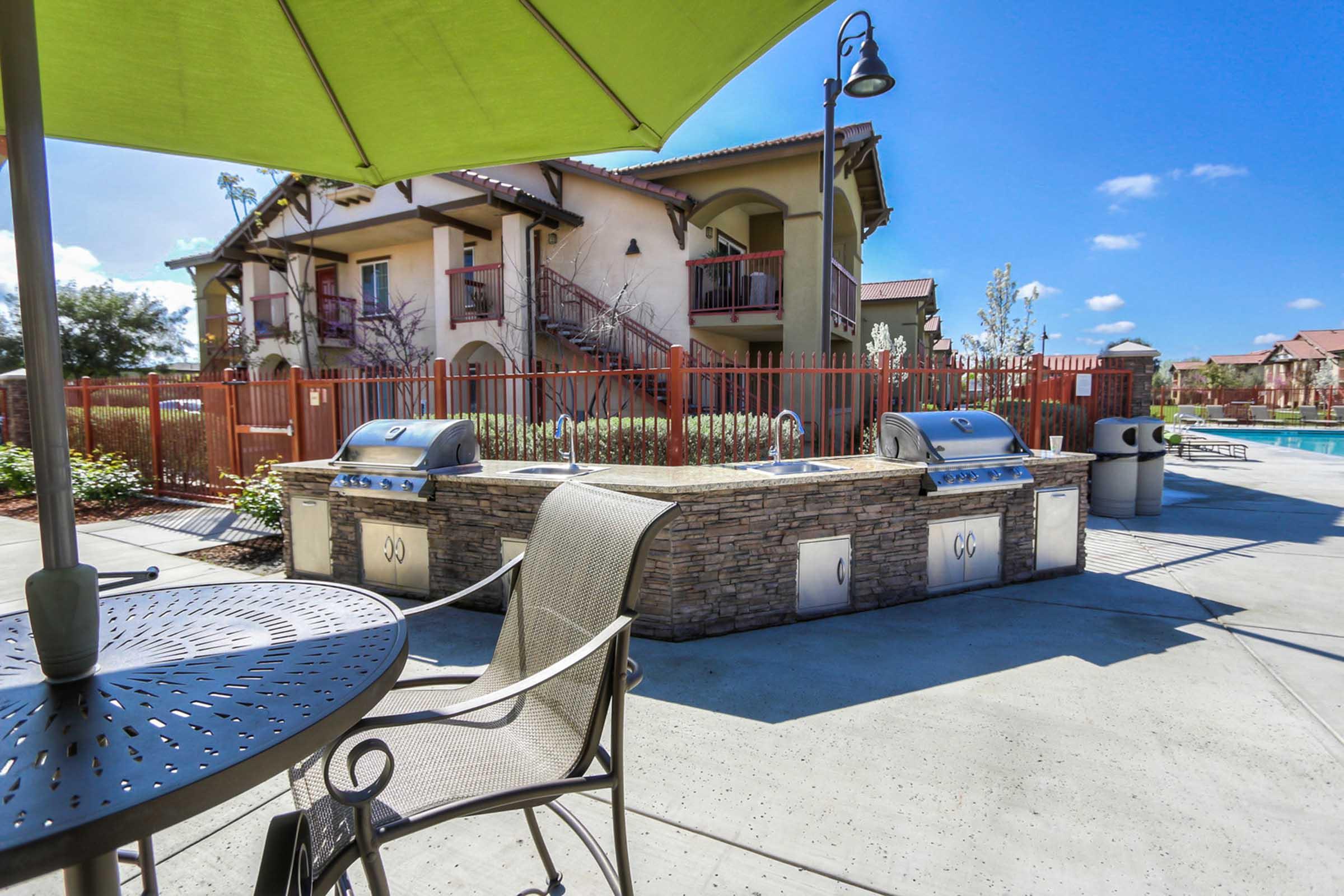 Outdoor barbecue area with two stainless steel grills, surrounded by stone counter space. Nearby is a table with a chair under a green umbrella. In the background, there are residential buildings with balconies and a clear blue sky. The area is well-maintained with landscaping.