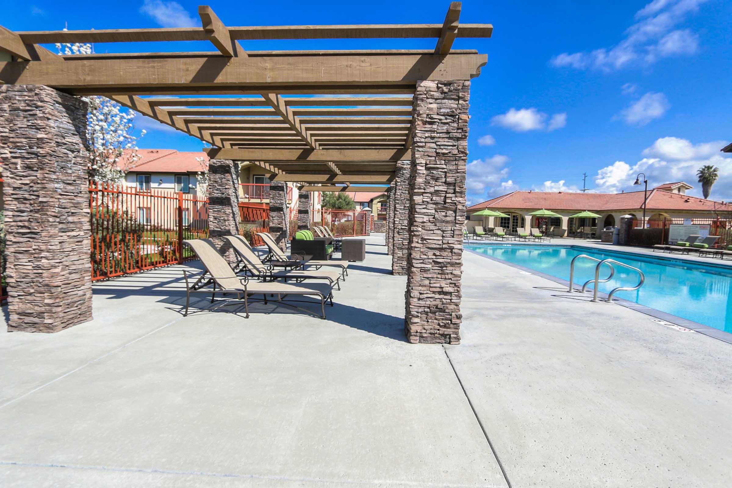 A sunny outdoor pool area with a stone pergola, featuring lounge chairs lined up on the concrete deck. The pool is calm and inviting, surrounded by greenery and residential buildings in the background under a clear blue sky with scattered clouds.