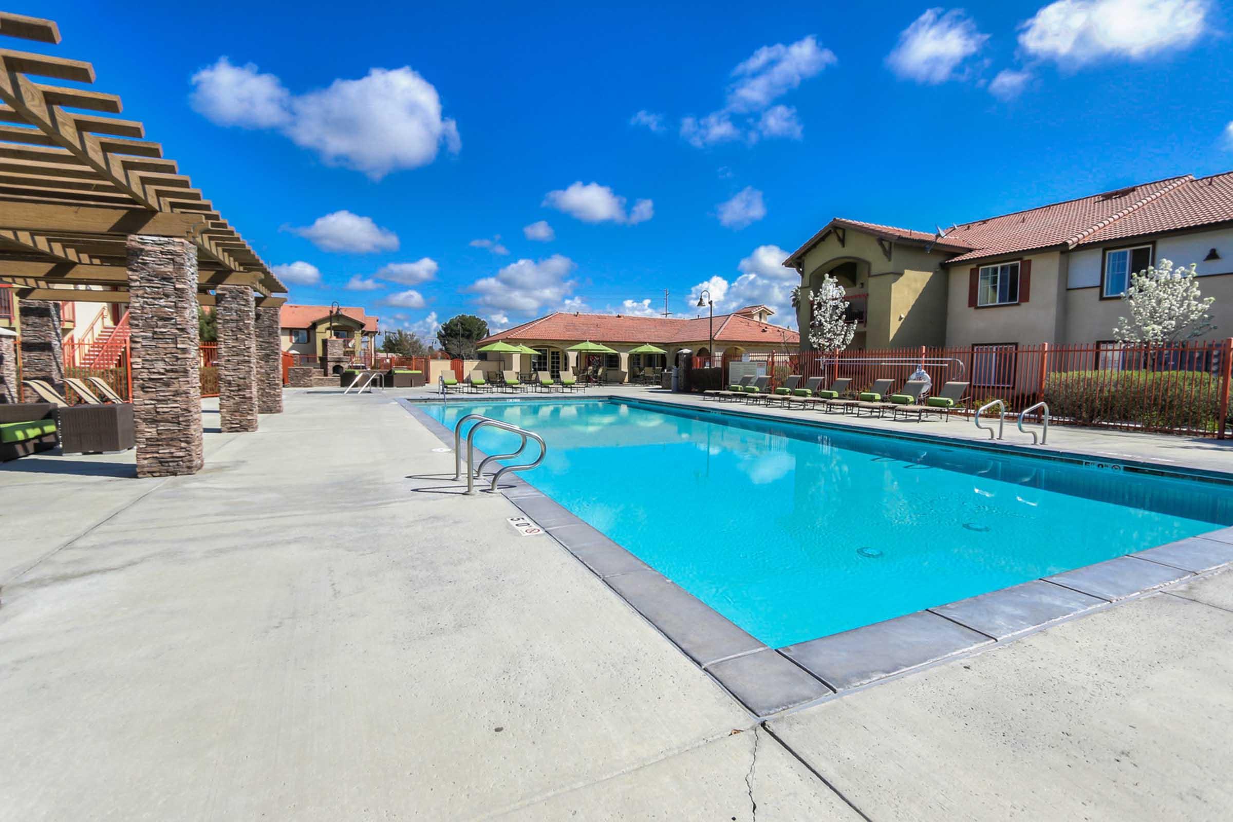 A view of a swimming pool surrounded by lounge chairs and shaded areas. In the background, there are residential buildings with red roofs and landscaping. The sky is clear with a few fluffy clouds, creating a bright and inviting atmosphere.