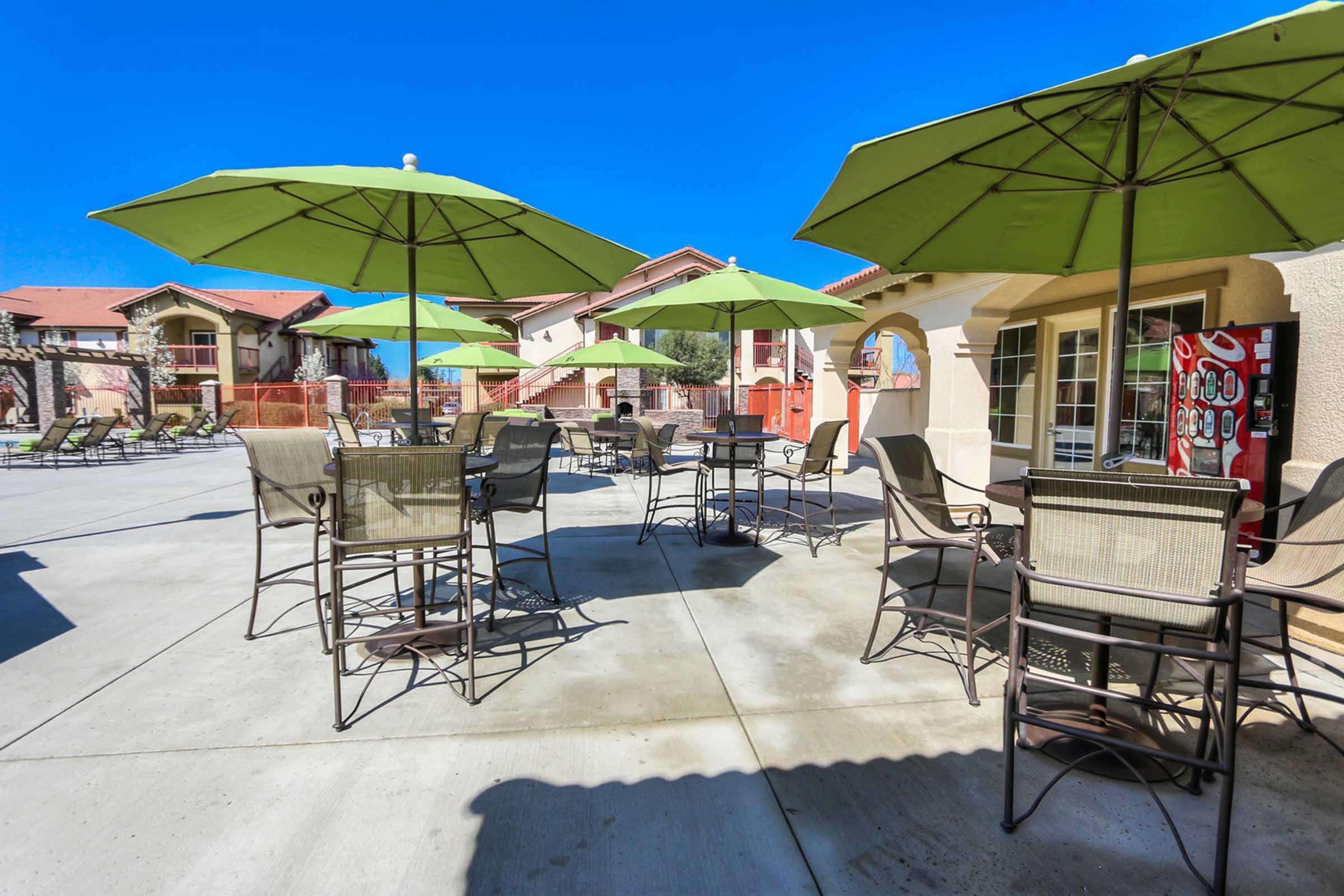 A spacious outdoor patio area with several tables and chairs shaded by green umbrellas. The ground is paved, and there are apartment buildings visible in the background. A soda vending machine is on one side of the patio, under a clear blue sky.