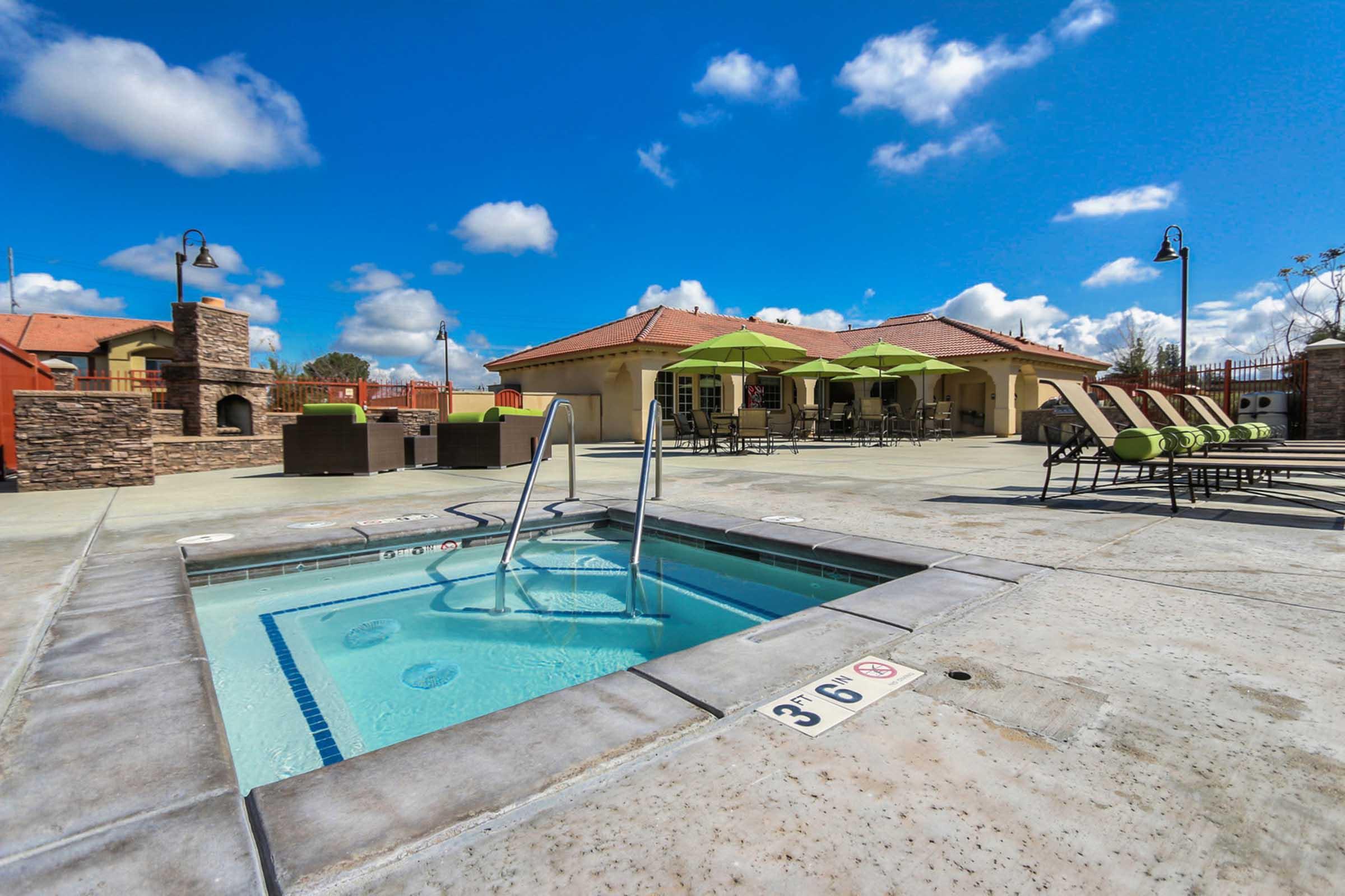A bright outdoor area featuring a hot tub with steps for entry, surrounded by lounge chairs and green umbrellas. In the background, there are stone structures and a building with a red roof under a blue sky with fluffy white clouds.