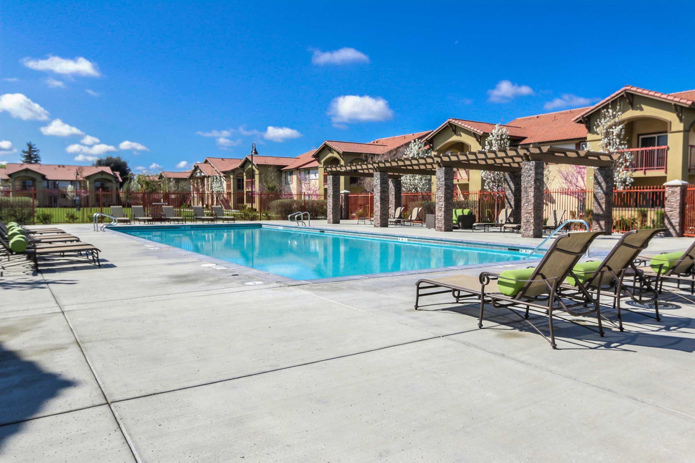 A sunny outdoor swimming pool area with a clear blue sky, surrounded by lounge chairs and shaded pergolas. In the background, there are residential buildings with red roofs and green grass. The scene conveys a relaxing atmosphere perfect for leisure and leisure activities.