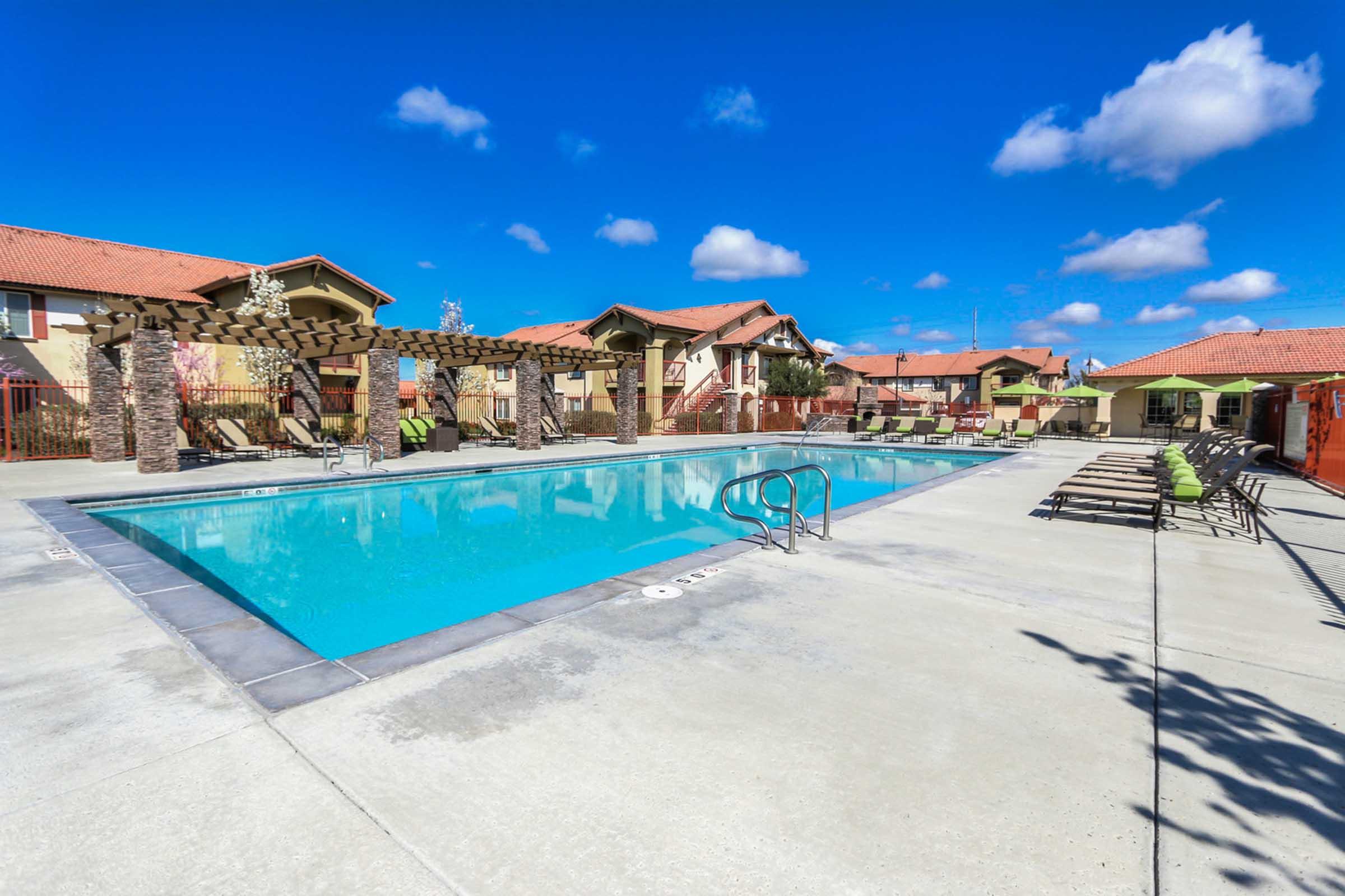 A clear, blue swimming pool surrounded by lounge chairs and shaded pergolas. In the background, there are residential buildings with orange roofs under a bright blue sky with scattered clouds. The area appears inviting and well-maintained, ideal for relaxation and leisure activities.
