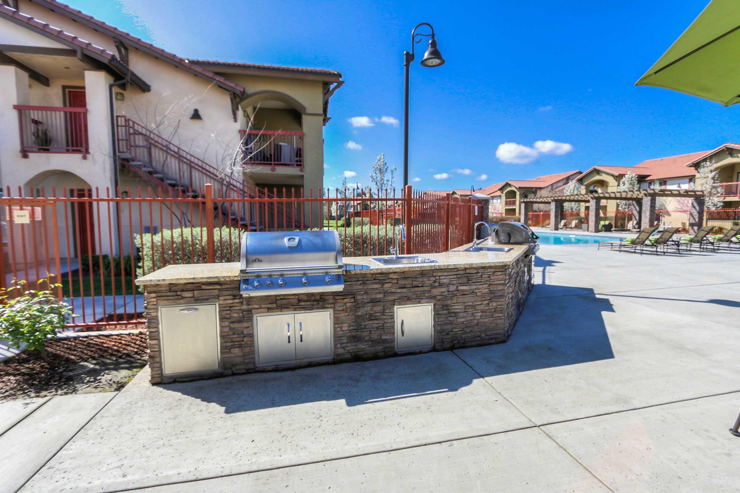 Outdoor grilling area with a stainless steel barbecue grill, stone countertop, and preparation space. Surrounding the area are red fences, residential buildings, a blue sky with scattered clouds, and a swimming pool visible in the background.