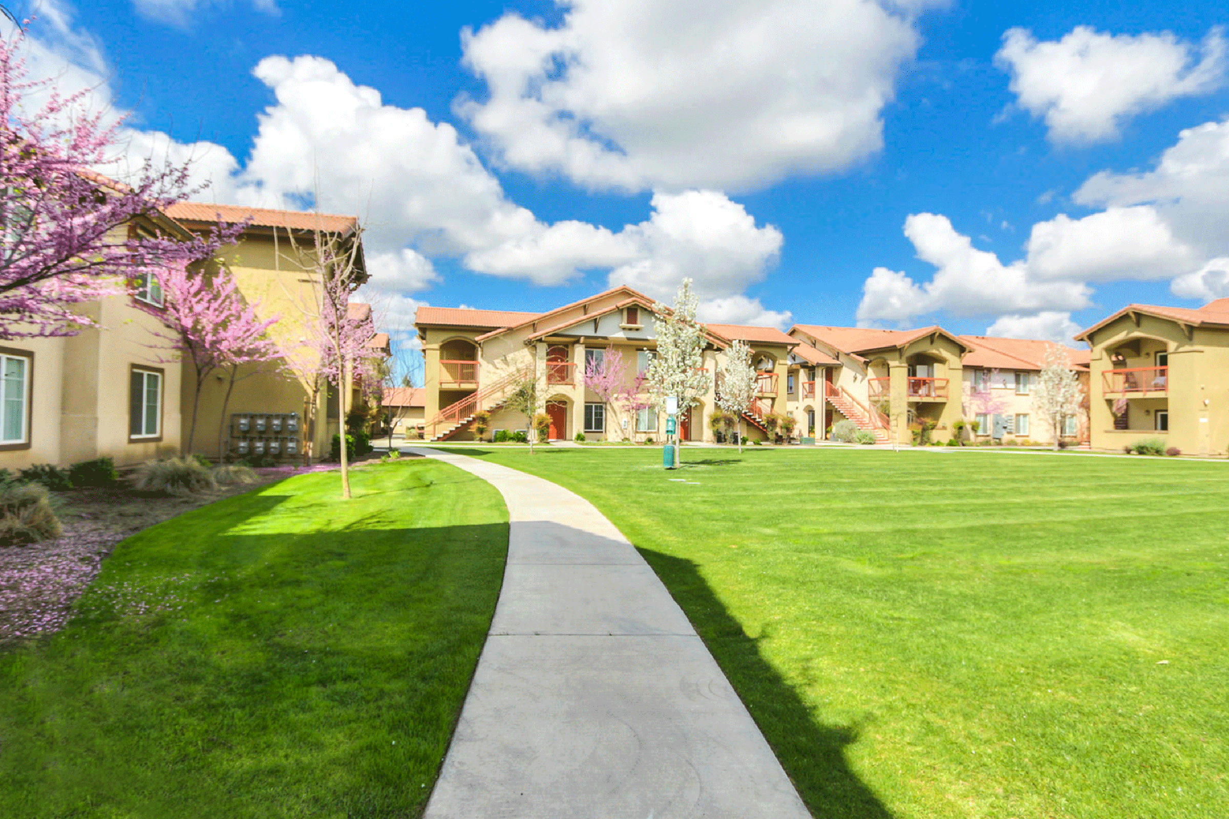 A scenic view of an apartment complex surrounded by green lawns and blooming trees under a bright blue sky with fluffy white clouds. A paved walkway meanders through the landscape, inviting residents to enjoy the outdoor space.