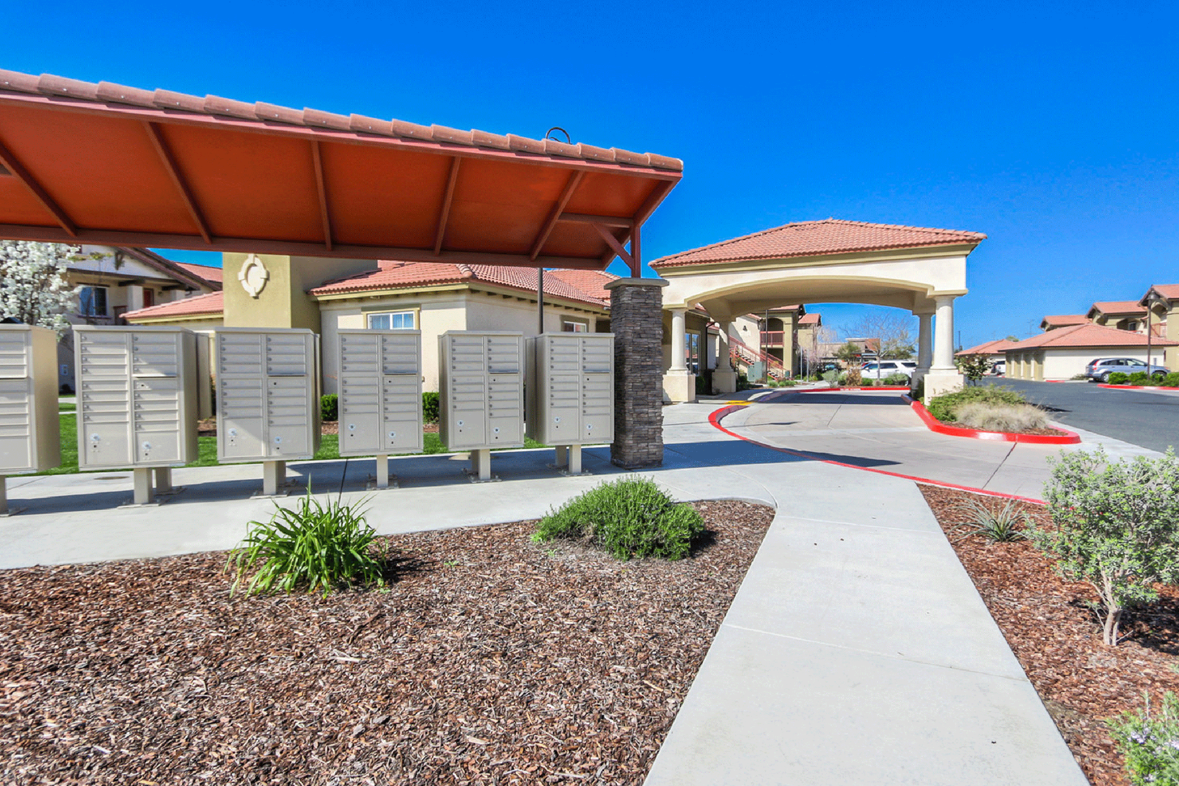 Gray residential mailboxes under a red-roofed structure, with a paved walkway leading to an entrance of an apartment complex. Lush green landscaping and a clear blue sky enhance the inviting atmosphere. The area appears well-maintained and residential.