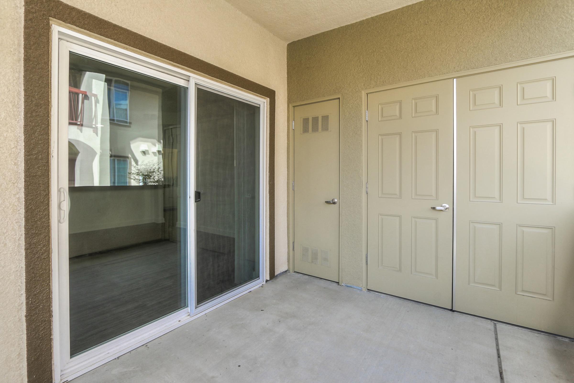 View of a small patio area featuring a large sliding glass door, providing access to an indoor space, alongside two closed tan doors on the right side. The concrete floor and beige walls create a neutral backdrop, suggesting a modern, minimalist design.
