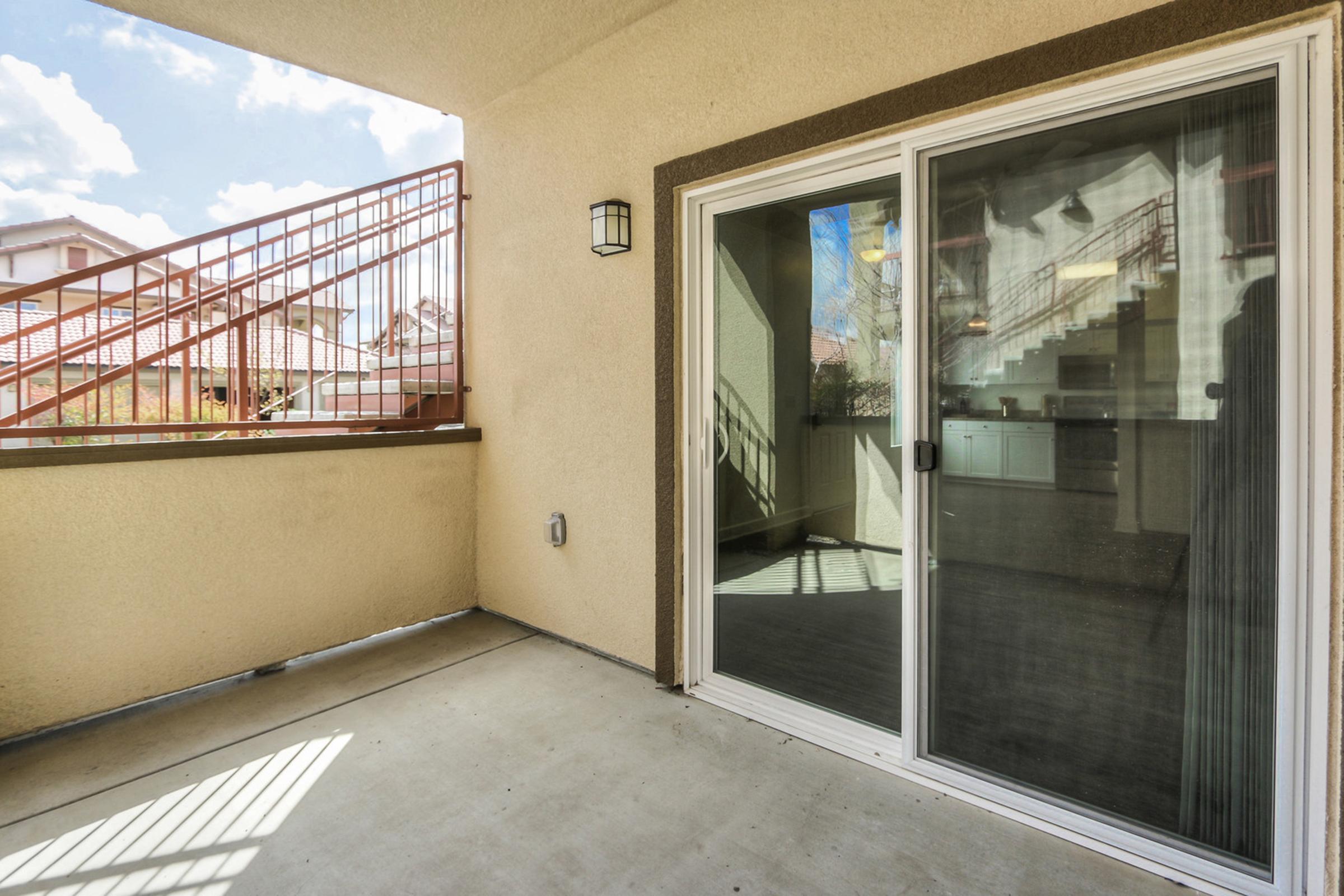 A spacious patio area featuring a sliding glass door that opens to a modern interior space. The wall is painted a light color, and there is a warm, natural light coming in through the door. A staircase is visible in the background, leading to another level of the building.