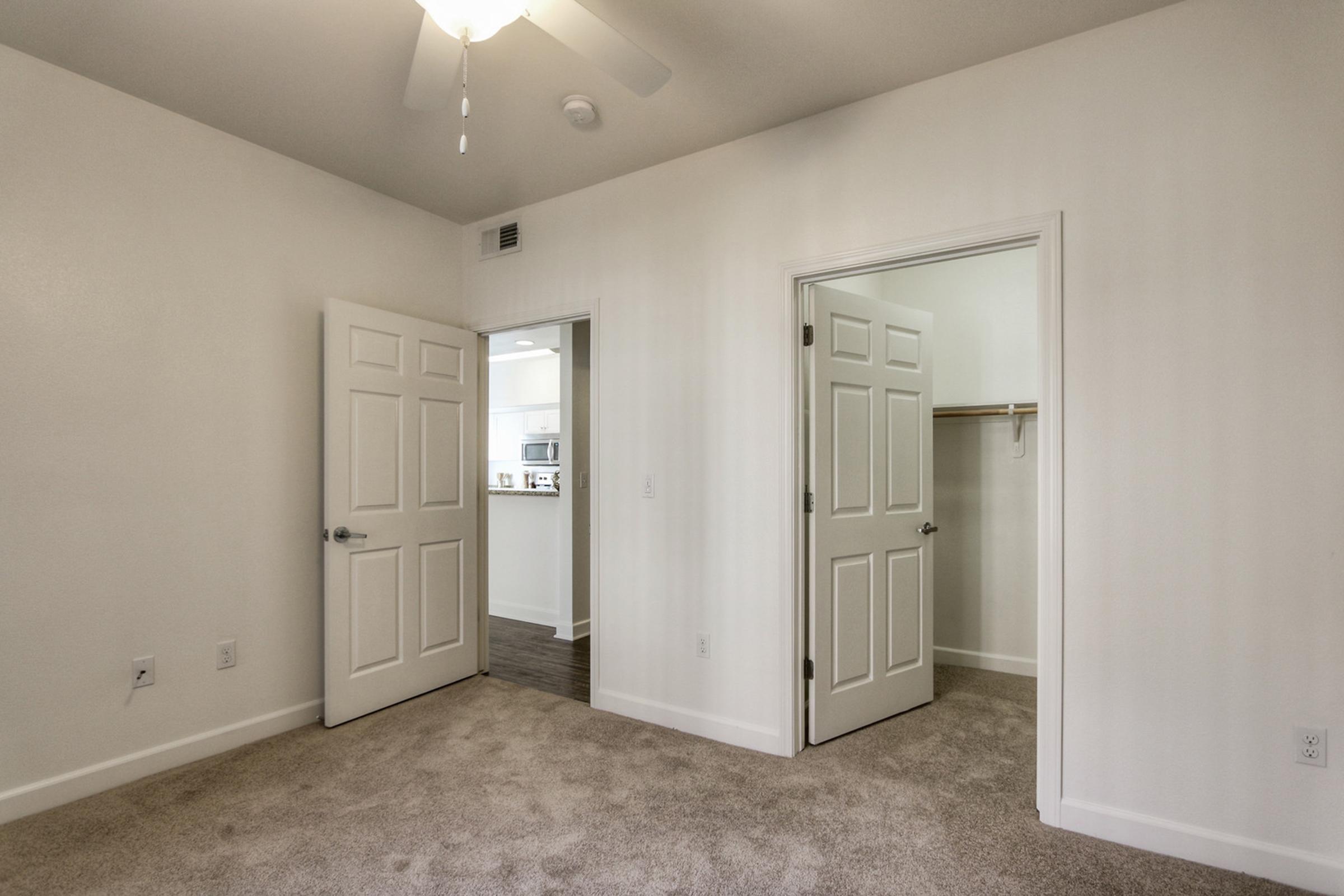 A spacious room with light-colored walls and carpeted flooring. There are two closed doors, one leading to a closet and the other to another room. A ceiling fan is visible above, adding to the airy feel of the space. The overall atmosphere is bright and inviting.