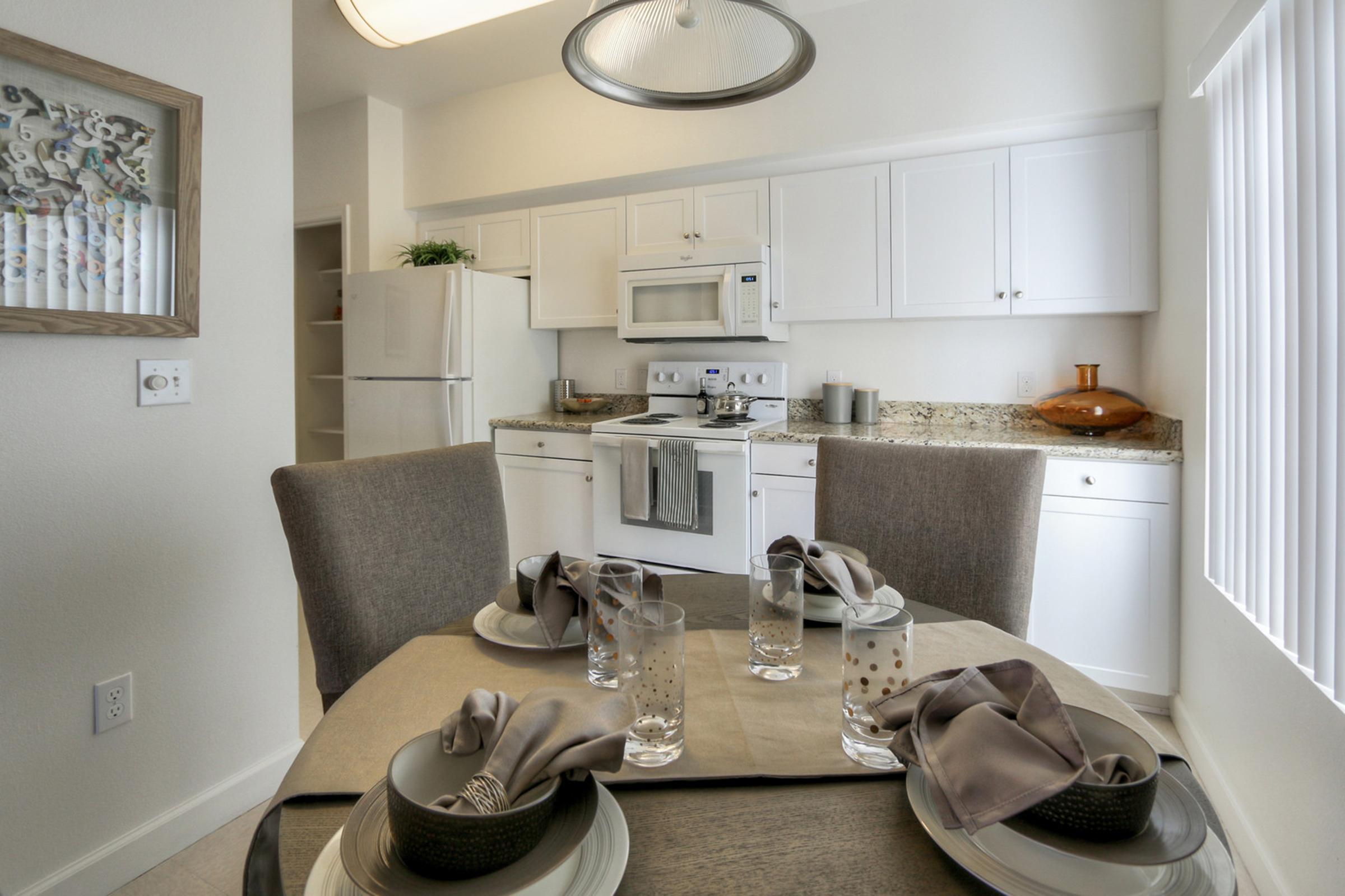 A modern kitchen with white cabinets and a granite countertop. A dining table is set with dishes, cloth napkins, and glassware. The room features natural light coming through window blinds. A refrigerator and microwave are visible in the background, contributing to the contemporary style.