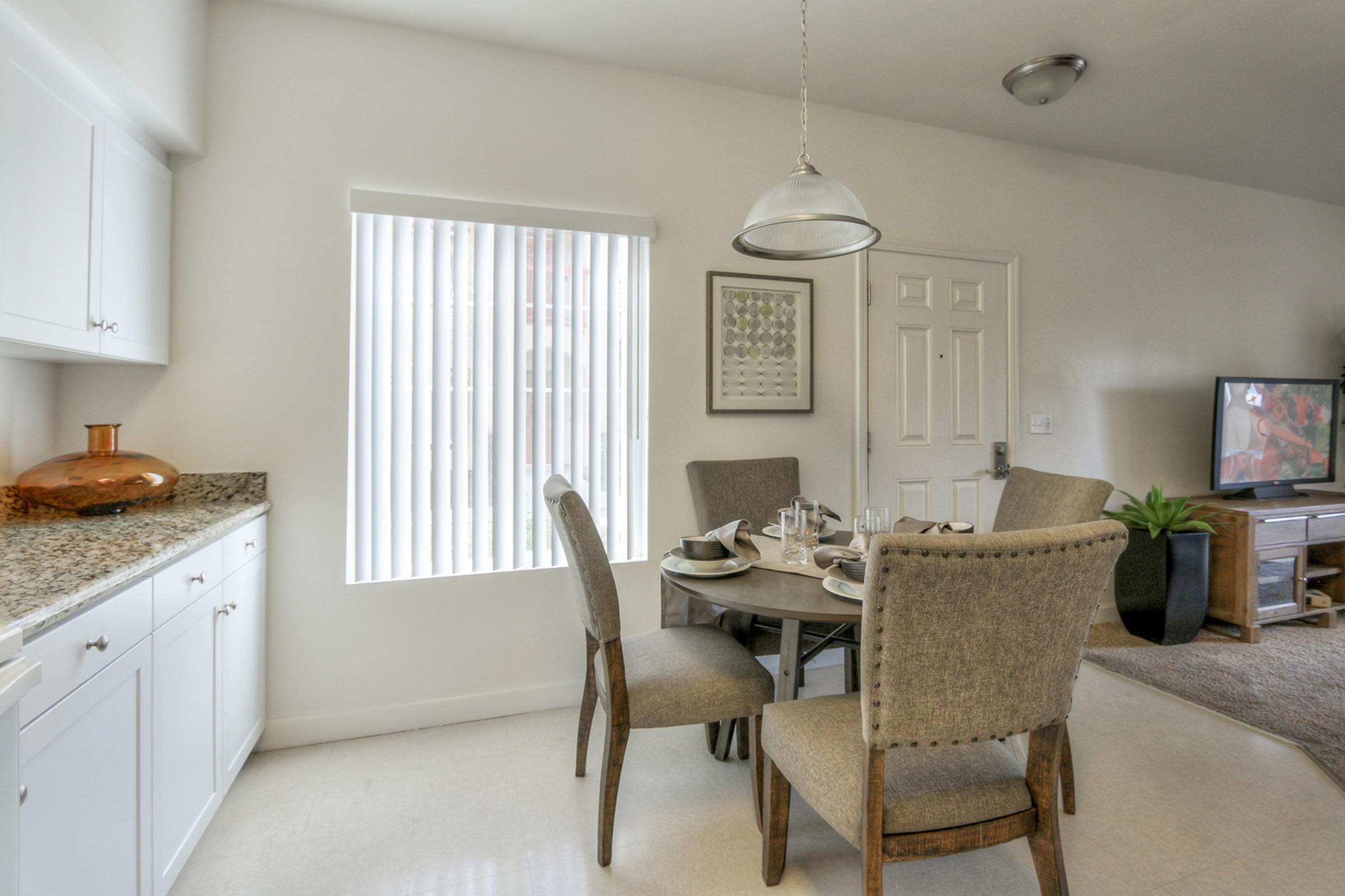 A modern dining area featuring a circular table set with dishes and cutlery, surrounded by four upholstered chairs. The kitchen has white cabinetry with a granite countertop and a decorative bowl. A window with vertical blinds allows natural light, and a television is visible in the corner of the room.