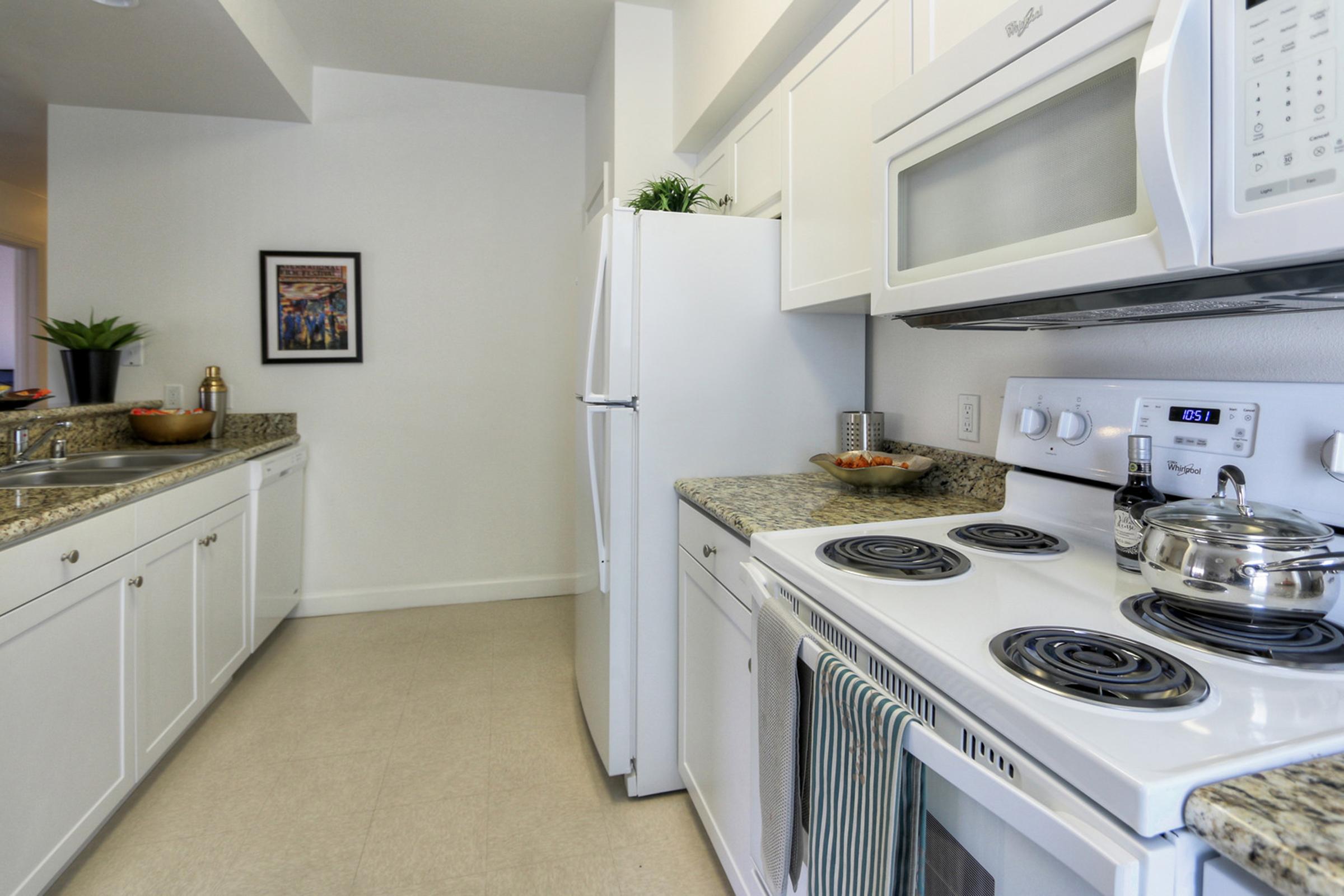A modern kitchen featuring white cabinetry, a granite countertop, a stove with four burners, and an oven. There is a microwave above the stove, a refrigerator on the left, and a sink with a bowl of fruit. The wall has a framed picture, and the room is well-lit with neutral-colored walls and flooring.