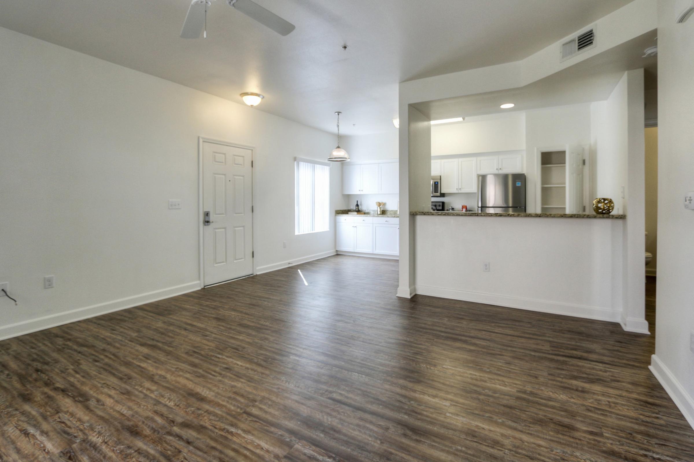 A spacious, modern living area with light wood flooring, featuring a white kitchen with stainless steel appliances visible in the background. Natural light streams in from a large window, and the room includes a ceiling fan and contemporary lighting fixtures. The layout is open and inviting.