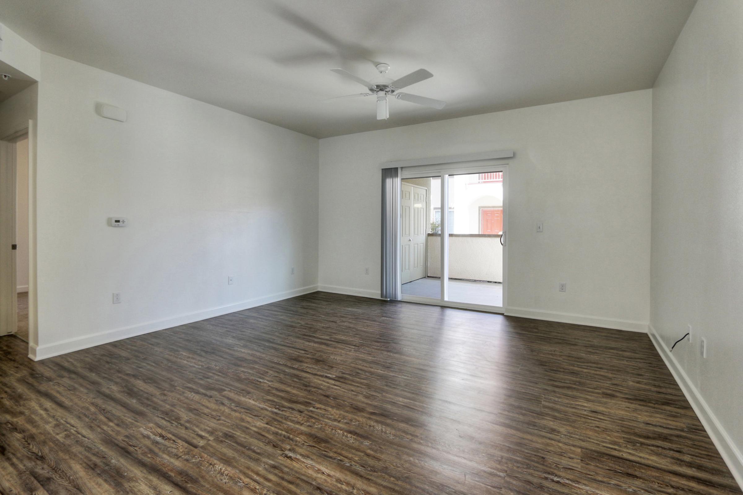 Interior of a spacious, empty living room with light-colored walls and a wood laminate floor. A ceiling fan is visible, and there is a sliding glass door leading to a small balcony. Natural light enters the room, creating a bright atmosphere.