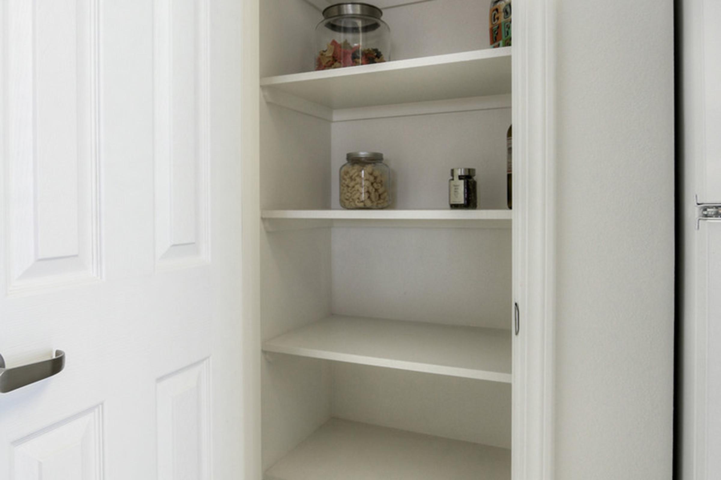 A white pantry cabinet with several empty shelves. A glass jar with a lid containing snacks is on the top shelf, alongside another container. The cabinet door is partially open, revealing the organized interior space.