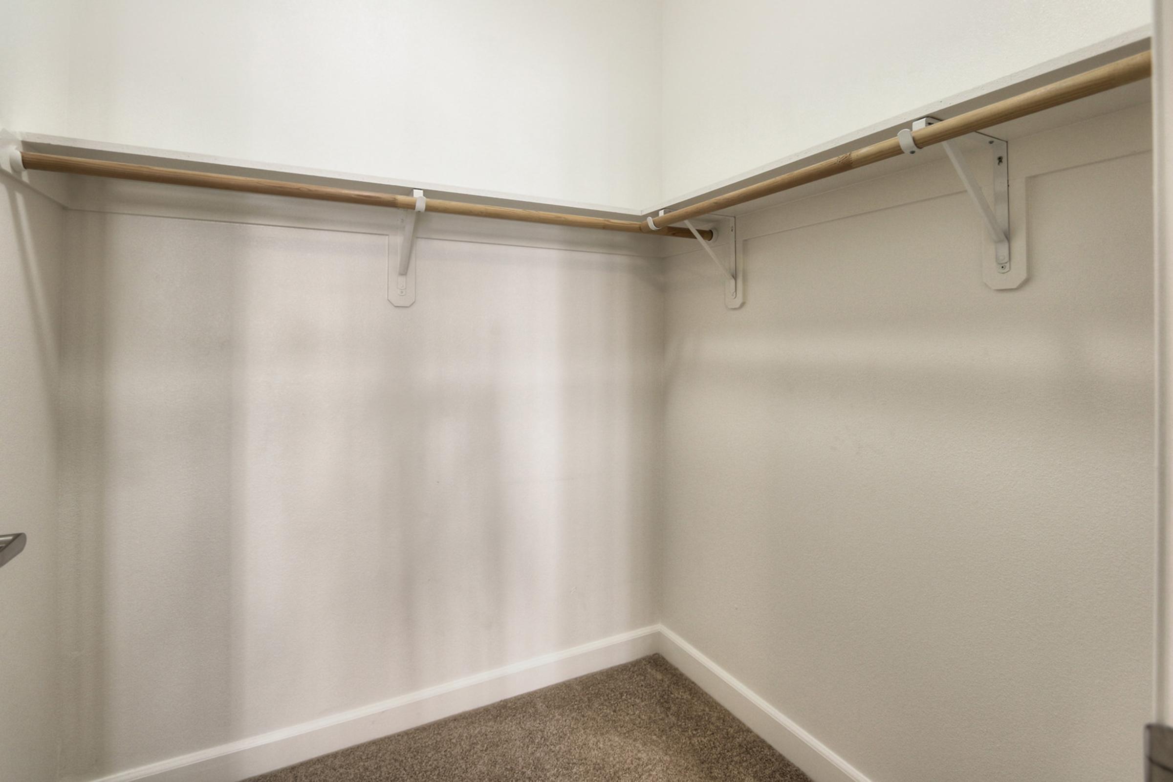 A spacious, empty closet featuring a light-colored wall and a hanging rod supported by white brackets. The floor is carpeted in a neutral tone, enhancing the bright and clean atmosphere of the space.