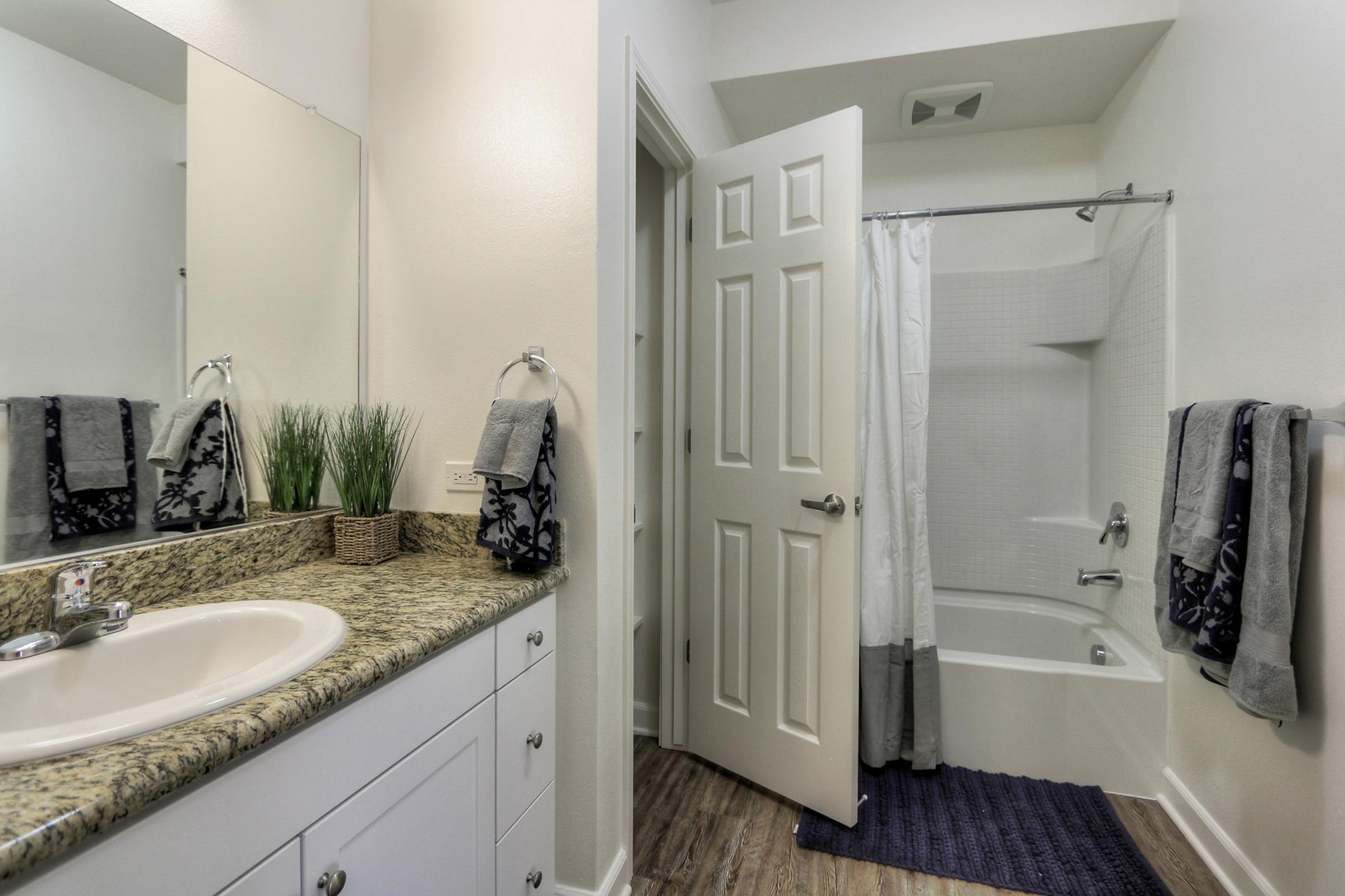 A modern bathroom featuring a light-colored countertop with a sink, white cabinets, and gray towels hanging on the wall. There is a shower/tub combination with a white curtain and a plant in a decorative pot. The door is partially open, revealing additional space in the bathroom.