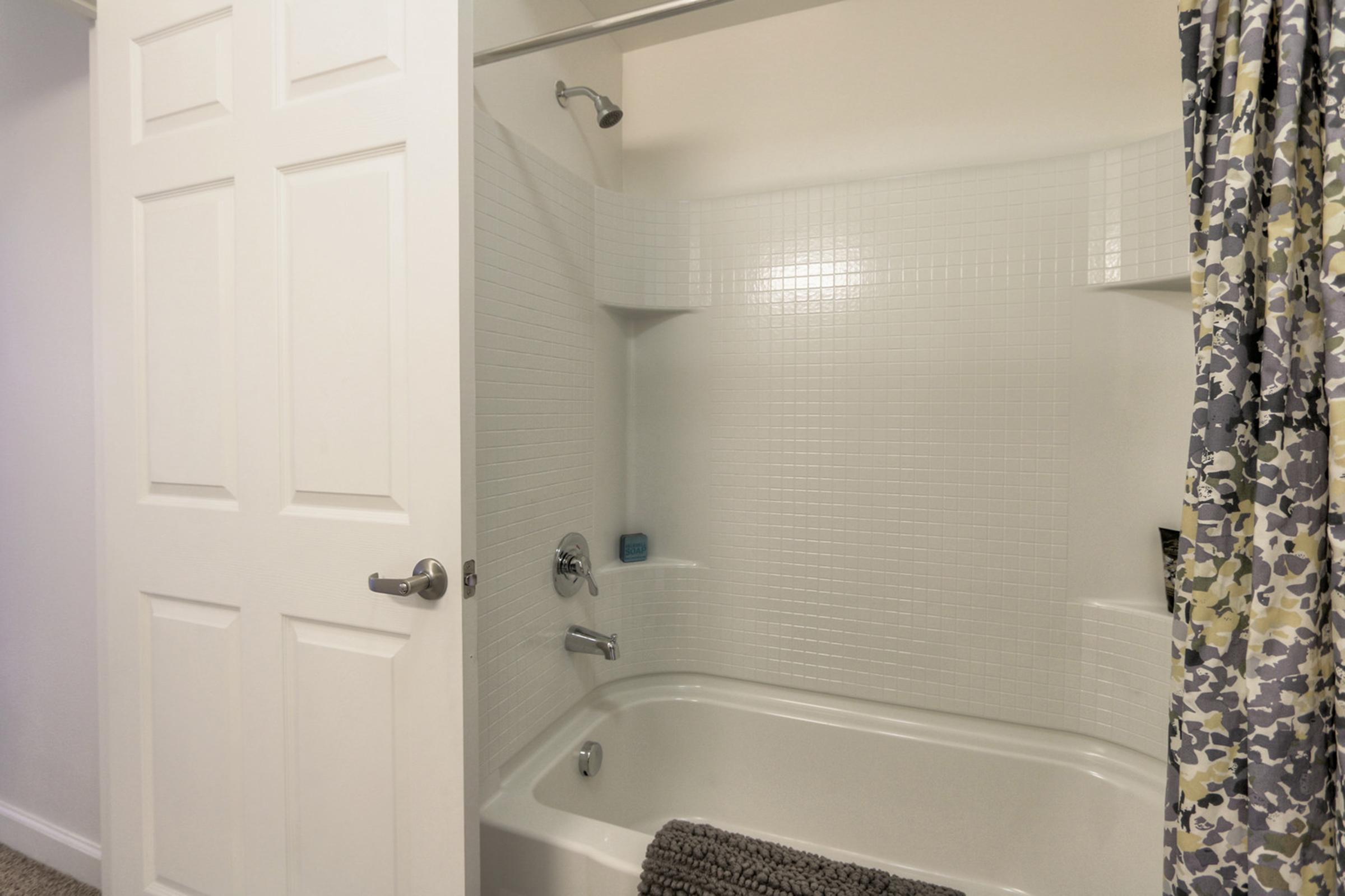 A clean bathroom interior featuring a white tiled shower and bathtub. The shower curtain has a colorful, patterned design, and there's a gray bath mat on the floor. A doorway is partially open, providing access to the bathroom. The lighting is bright and inviting, creating a fresh atmosphere.