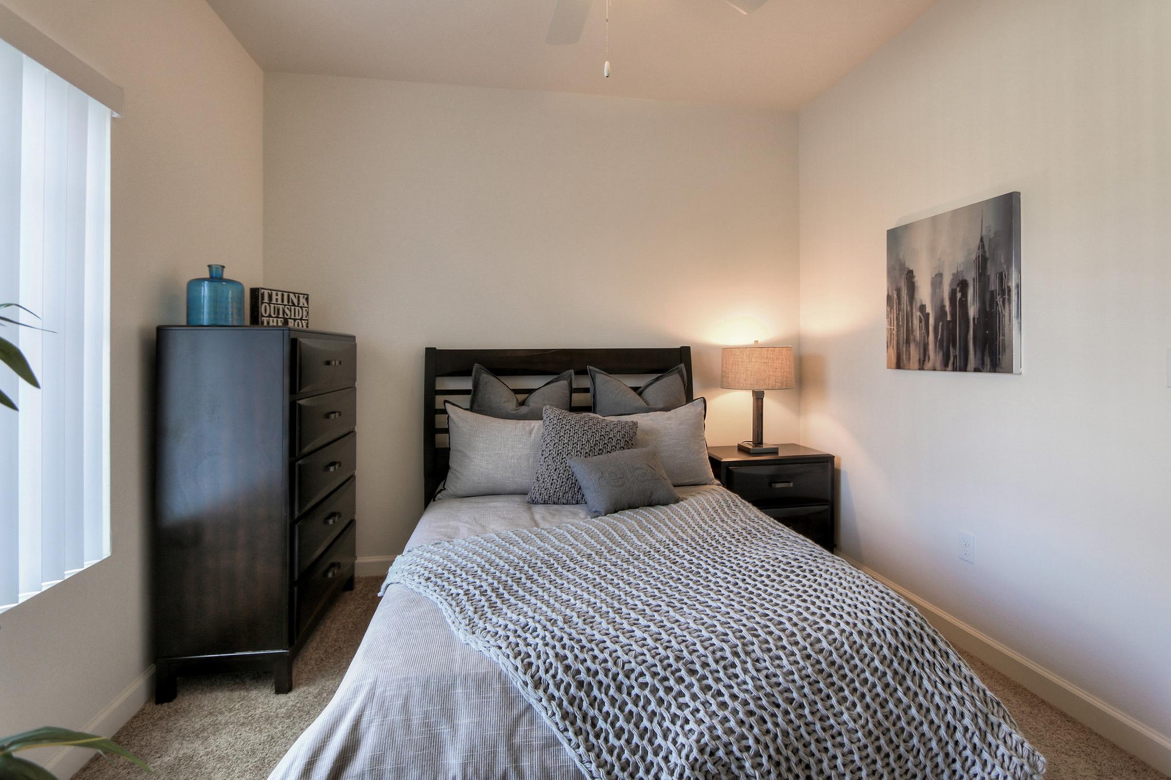 A cozy bedroom featuring a queen bed with decorative bedding, a dark wooden nightstand with a lamp, and a dresser. The walls are painted white, and there is a piece of cityscape artwork above the bed. Natural light enters through a window with blinds, creating a warm and inviting atmosphere.