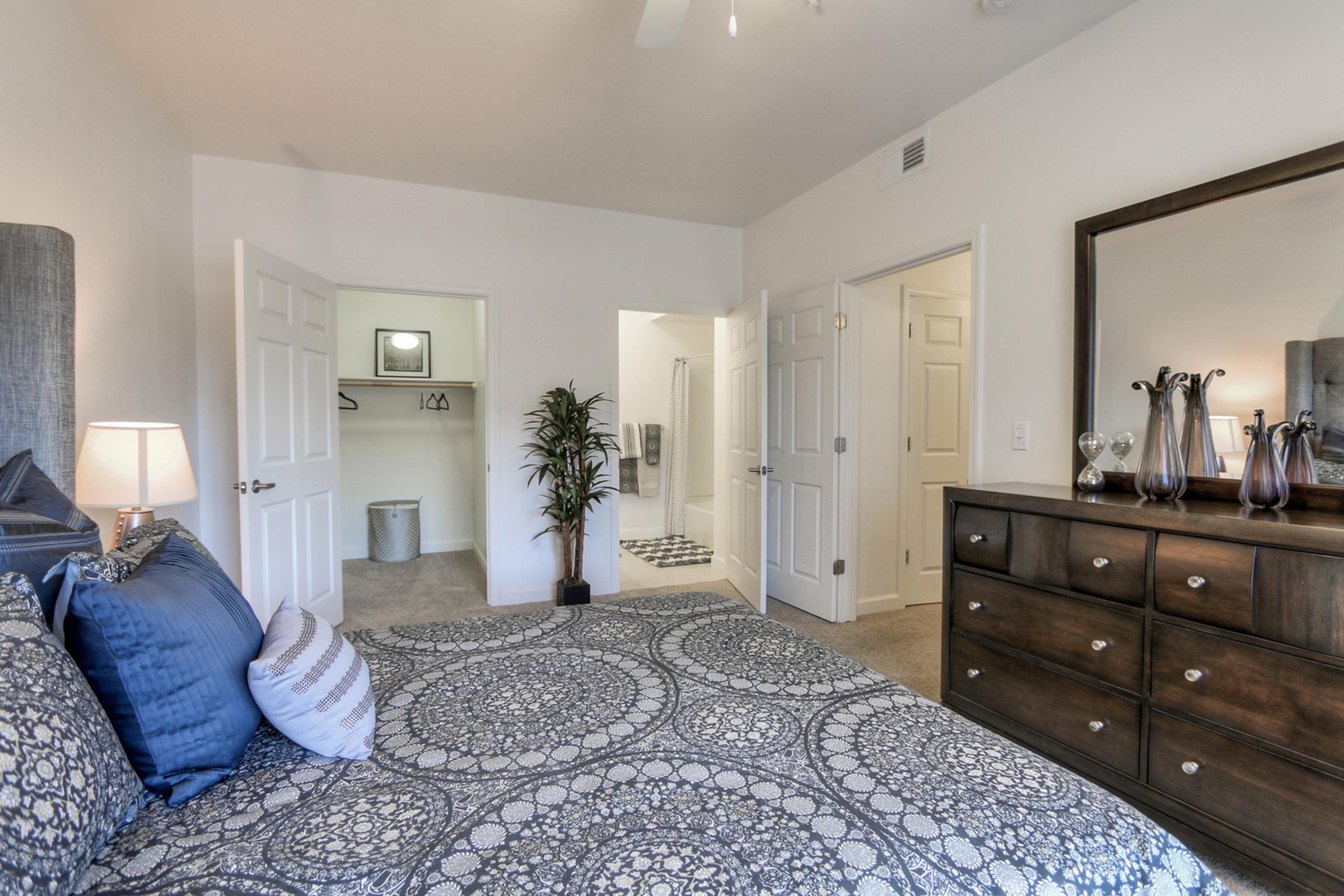 A cozy bedroom featuring a large bed with patterned bedding, a wooden dresser, and a decorative mirror. There are double doors leading to a closet and bathroom area. Soft lighting adds warmth, while a potted plant enhances the inviting atmosphere of the space.