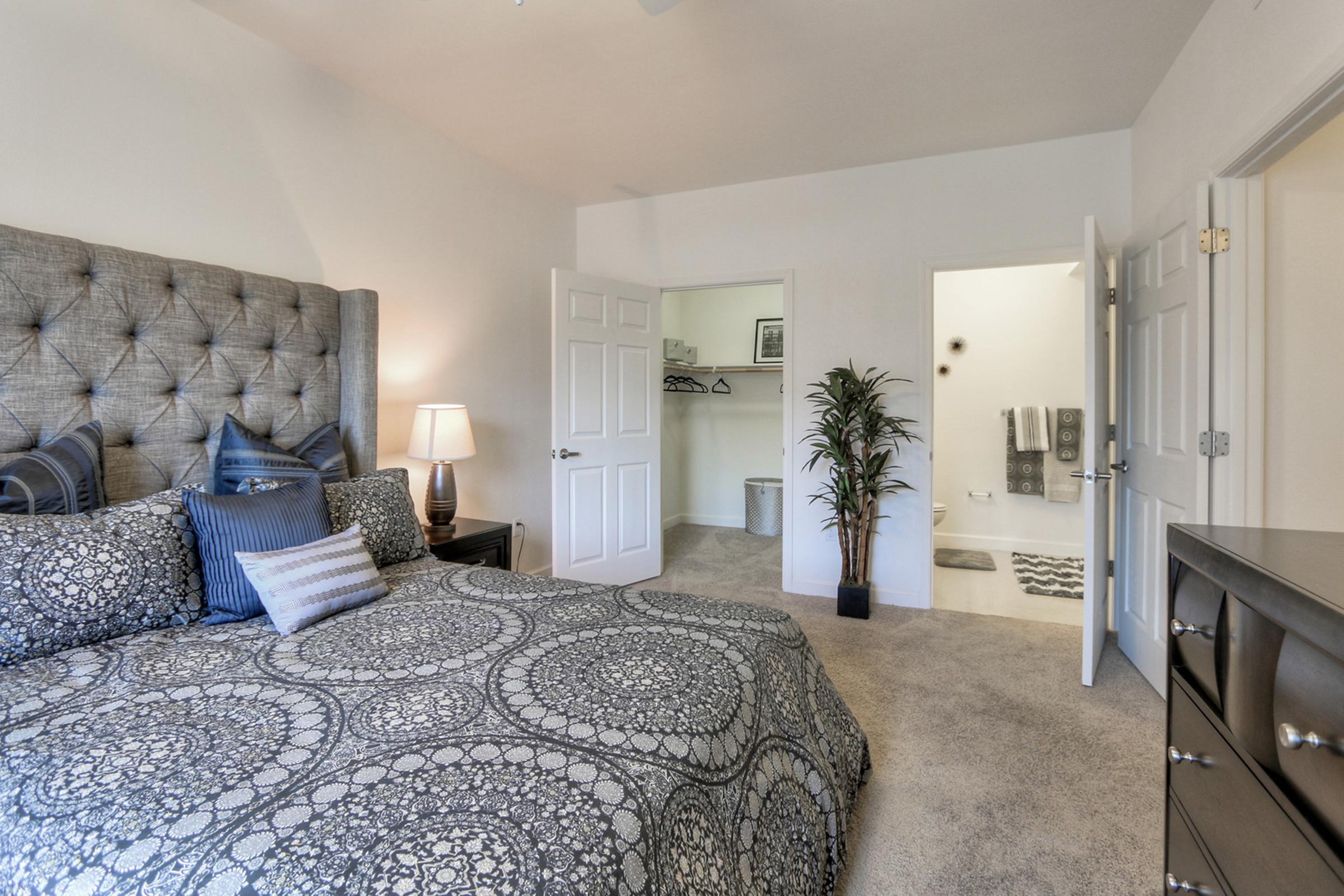 A cozy bedroom featuring a large tufted headboard with decorative pillows, a wooden nightstand with a lamp, and a plush carpet. The room connects to a bathroom area visible through an open door, which includes a towel rack and additional decor, creating a warm and inviting atmosphere.