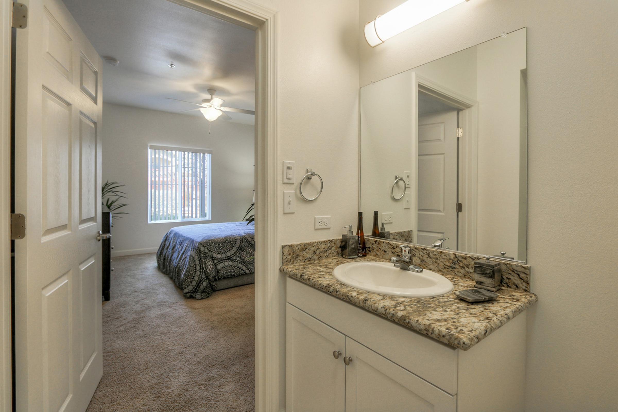A well-lit bathroom with a vanity featuring a sink and granite countertop. A mirror hangs above the sink, and there are towels and toiletries on the counter. In the background, a bedroom is visible with a bed covered in a patterned bedspread and a ceiling fan, adding a cozy ambiance to the space.