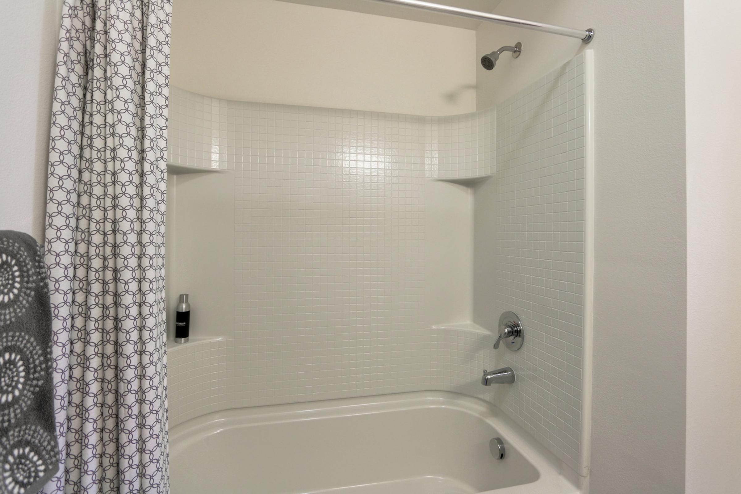 A clean, modern shower stall with white tile walls and a tub. There is a silver showerhead and a simple curtain with a geometric pattern. A bottle is placed on the edge of the tub, and the overall ambiance is bright and minimalist.