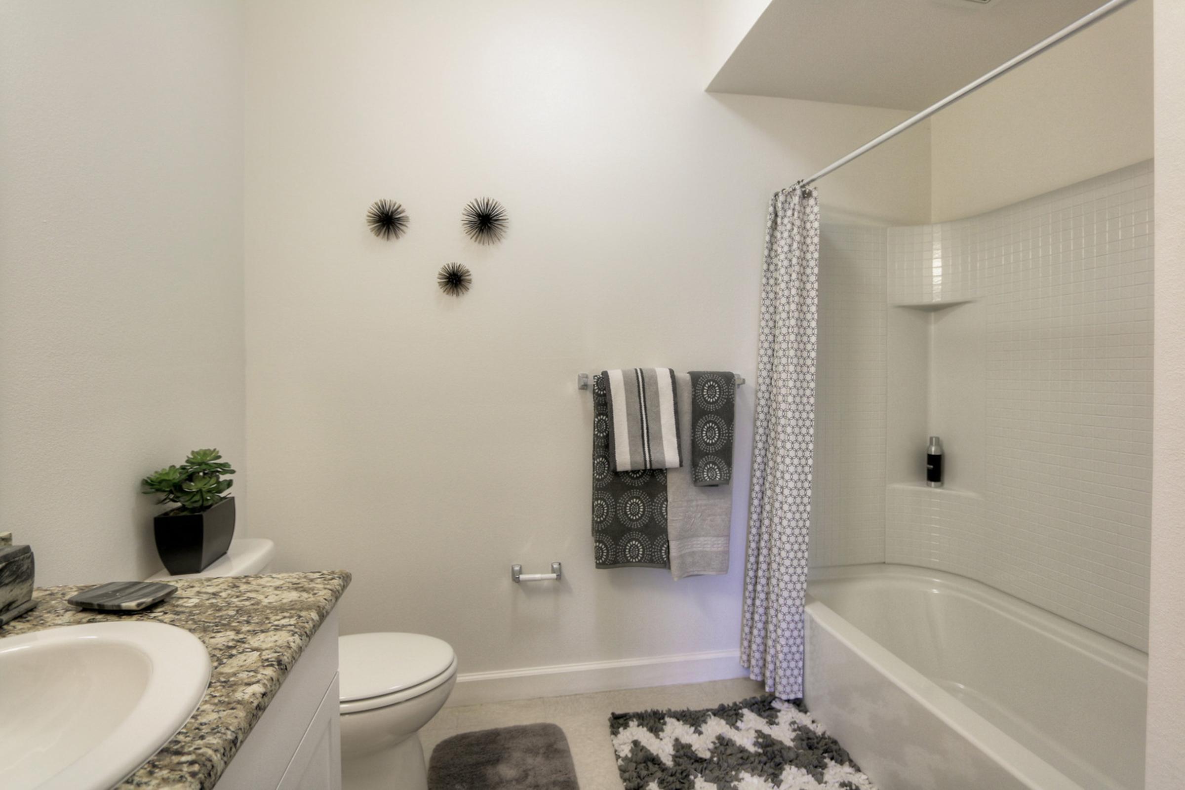 A bright and modern bathroom featuring a white tub/shower combo, light-colored walls, and a granite countertop. There are decorative towels, a small plant, and wall decor made of black spiky shapes. The space is neatly arranged with neutral colors and minimalistic design elements.