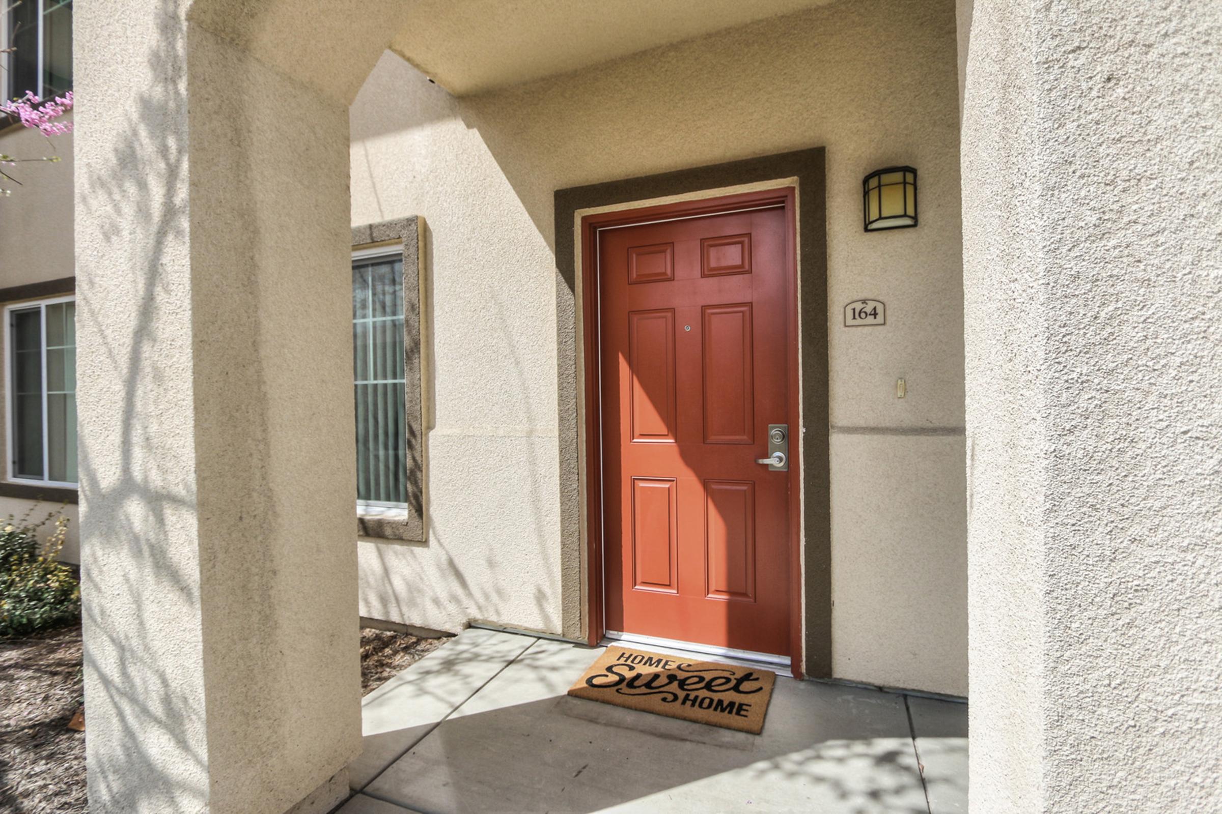 A brightly colored red door with a silver handle, set in a beige wall. A welcome mat with "Home Sweet Home" text is positioned in front of the door. The entrance area is framed by a small porch and features light-colored walls, plants, and a warm, inviting atmosphere.