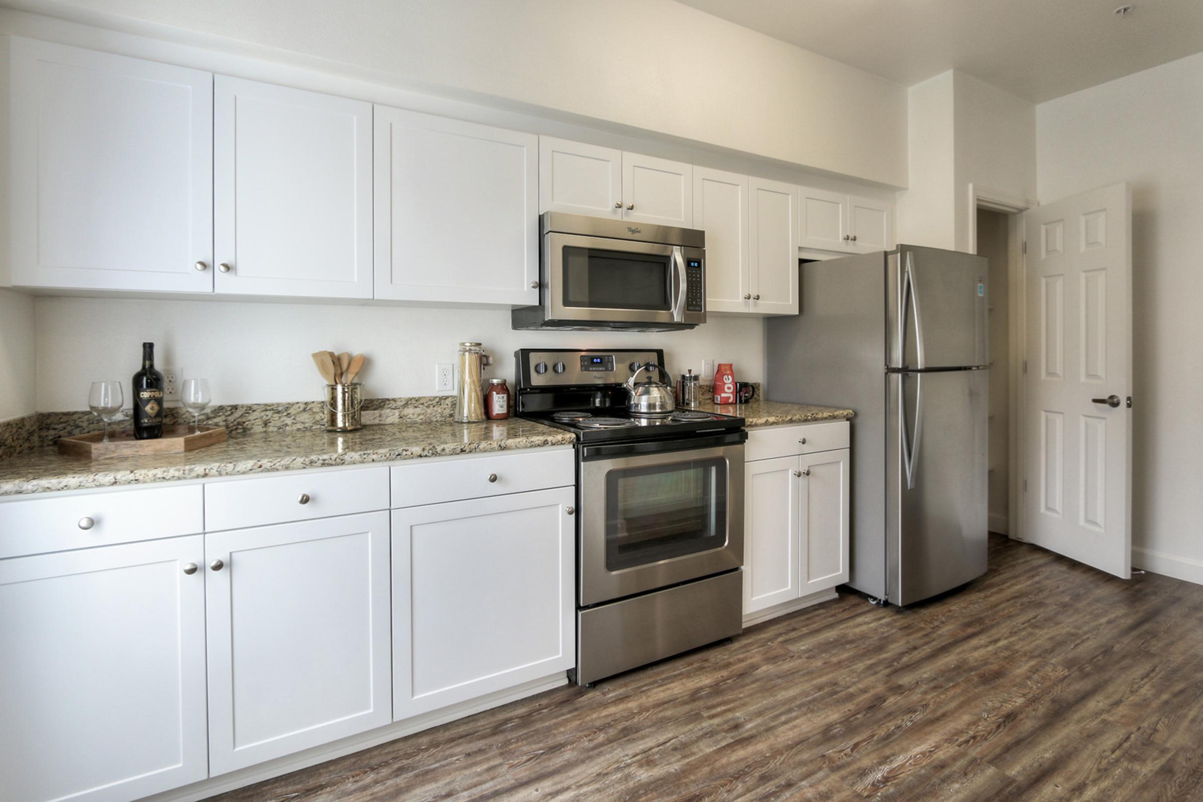 Modern kitchen with white cabinetry, stainless steel appliances including an oven and refrigerator, and a granite countertop. There are wine glasses on the counter, and the space has a light, airy feel with wooden floors and a partially open door leading to another room.