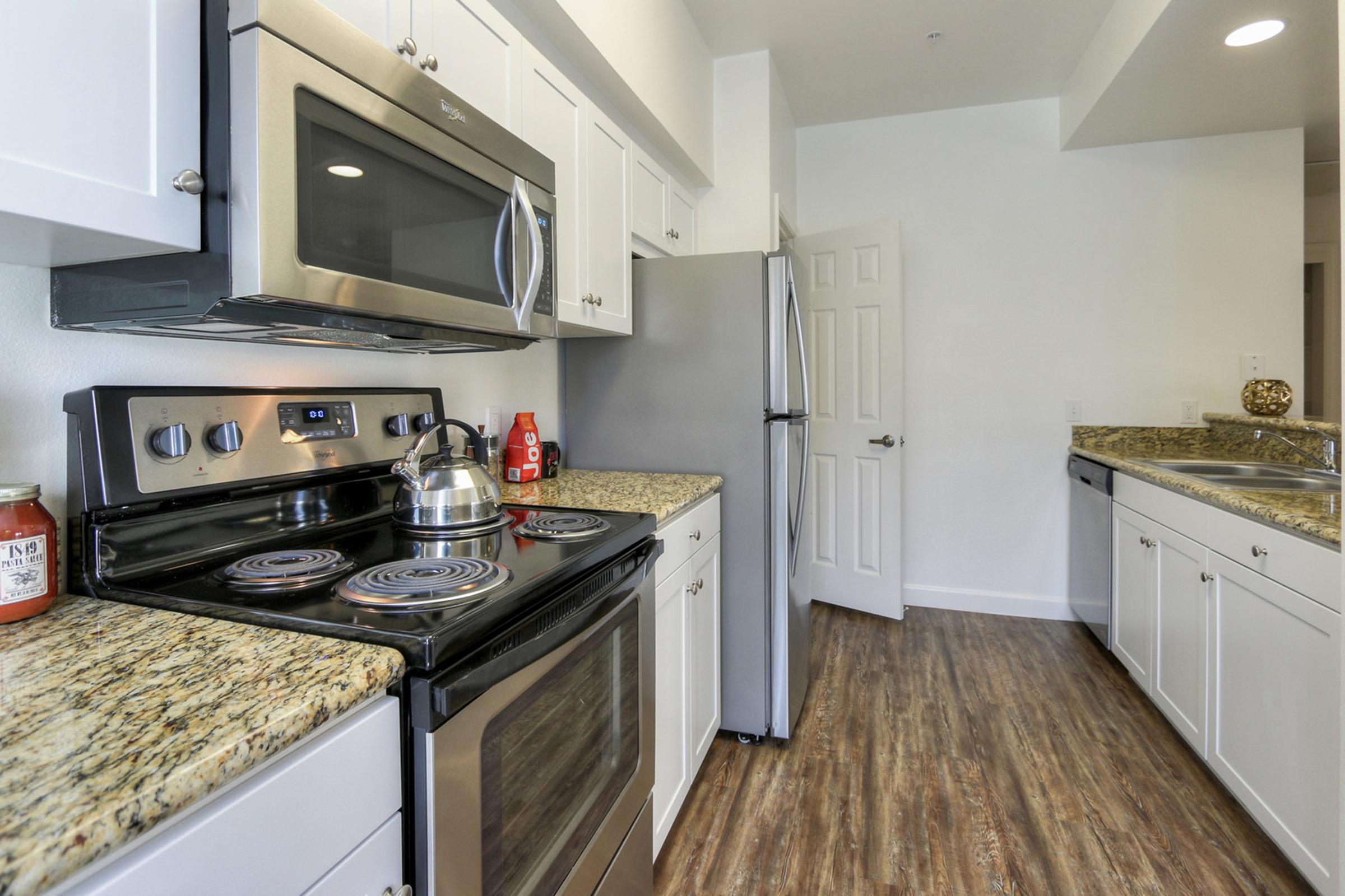 Modern kitchen featuring white cabinetry, stainless steel appliances, and granite countertops. An electric stove with a kettle on the burner, a microwave above, and a refrigerator on the right. The floor has a wood-like finish, and there is a doorway leading to another room visible in the background.
