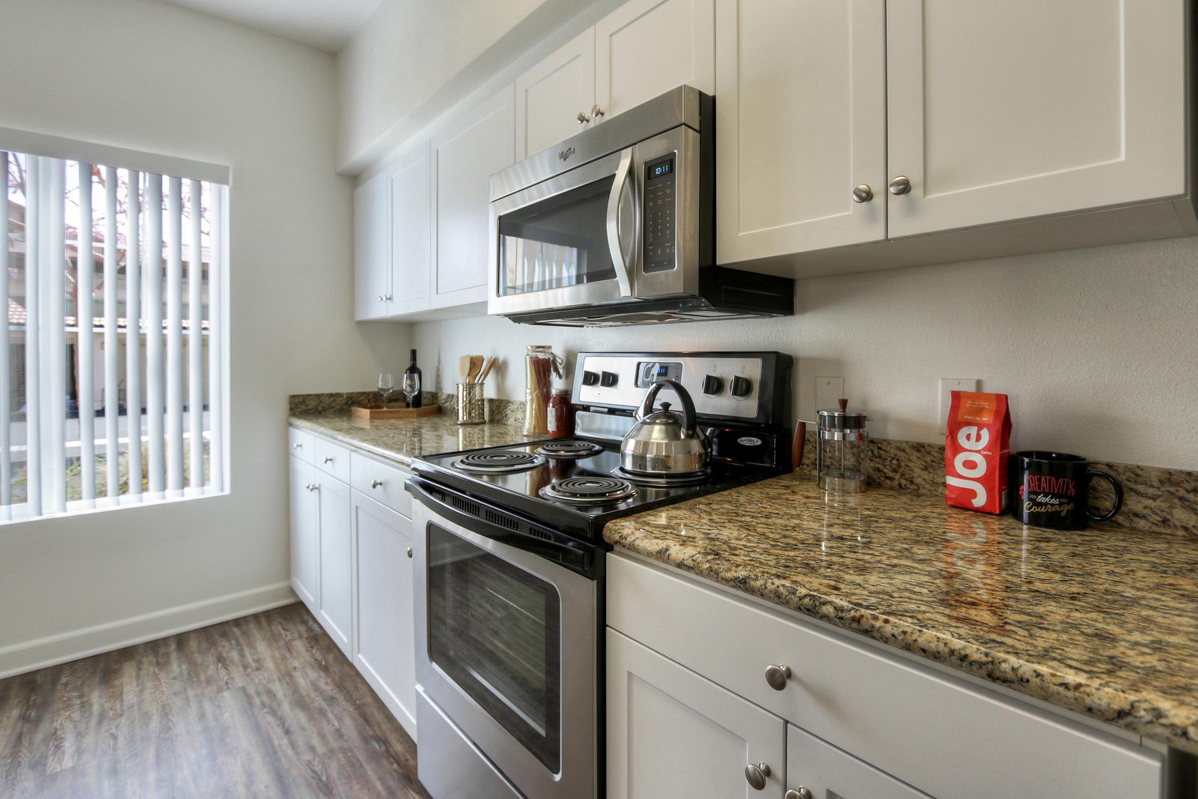 Modern kitchen with white cabinets, granite countertops, and stainless steel appliances. Features a stove, microwave, and kettle, with kitchen utensils and a coffee mug on the counter. A window with blinds provides natural light, enhancing the bright and inviting atmosphere.