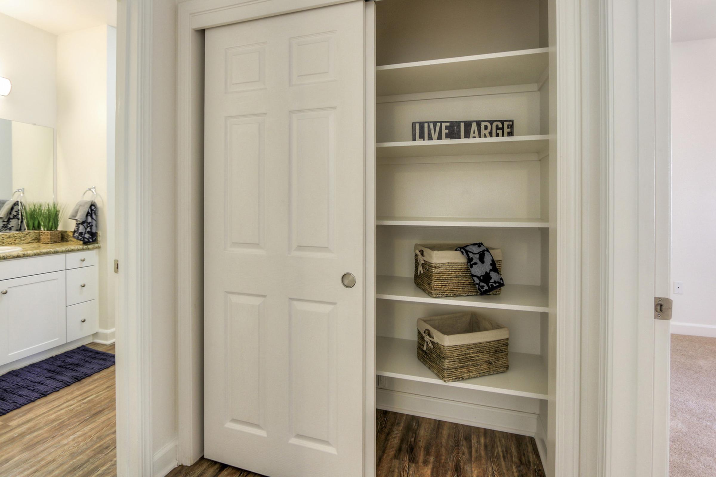 Interior view of a closet with white double doors slightly ajar. Inside, there are several empty shelves, one holding decorative items that read "LIVE LARGE." Baskets are placed on the shelves, and a small portion of a bathroom vanity is visible in the background, featuring a mirror and counter with plants.