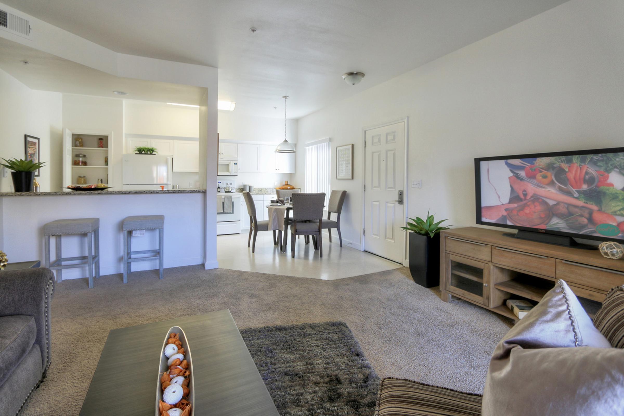 A cozy living room featuring a plush sofa, a decorative coffee table with a centerpiece, and a television. In the background, there is a dining area with a set of chairs and a kitchen visible. The space has neutral-colored walls and carpet, with houseplants adding a touch of greenery.