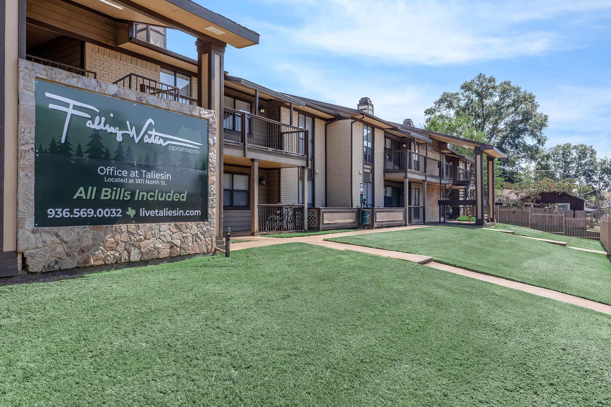 A well-maintained apartment community featuring a large sign for "Talleisin" advertising office contact information. The building has multiple balconies, manicured green lawns, and a clear blue sky in the background. The sign states "All Bills Included" and provides a phone number for inquiries.