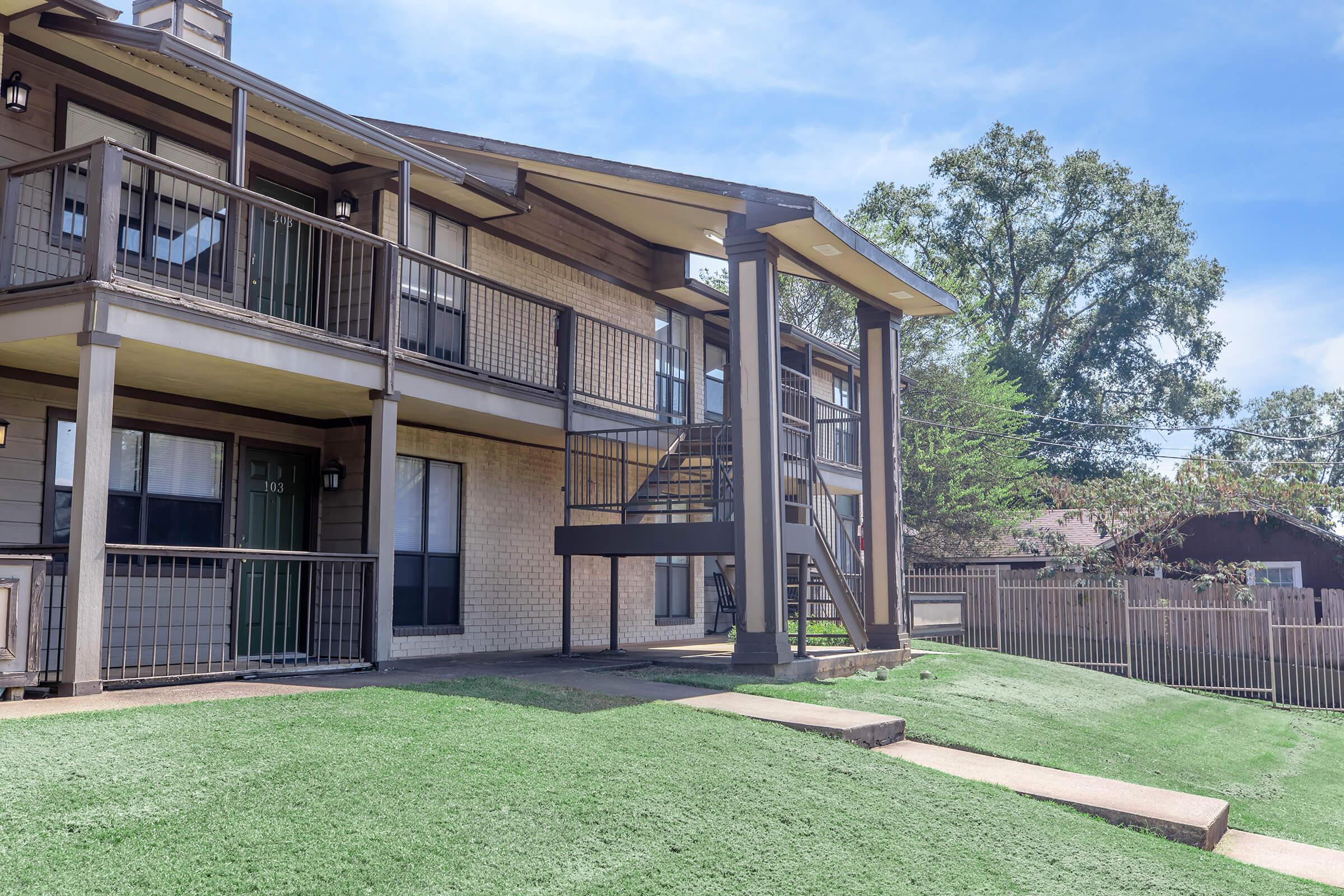 A three-story apartment building with balconies and a central staircase. The building features brick and siding exteriors, surrounded by green grass and a pathway. In the background, a fence and trees are visible under a clear blue sky.