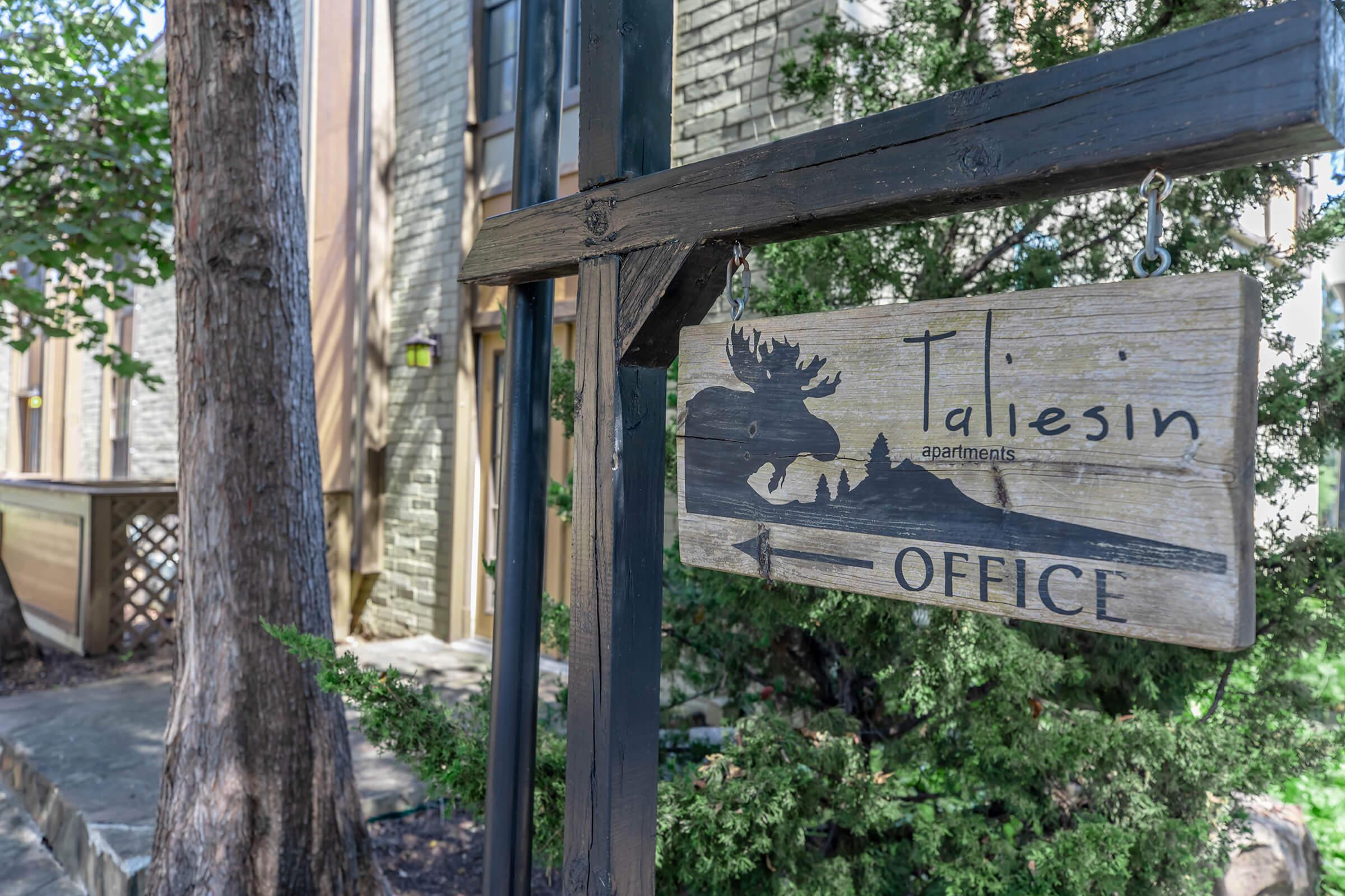 A wooden sign for "Taliesin Apartments" featuring an illustration of a moose, directing toward the office. The background includes part of the apartment building and greenery.